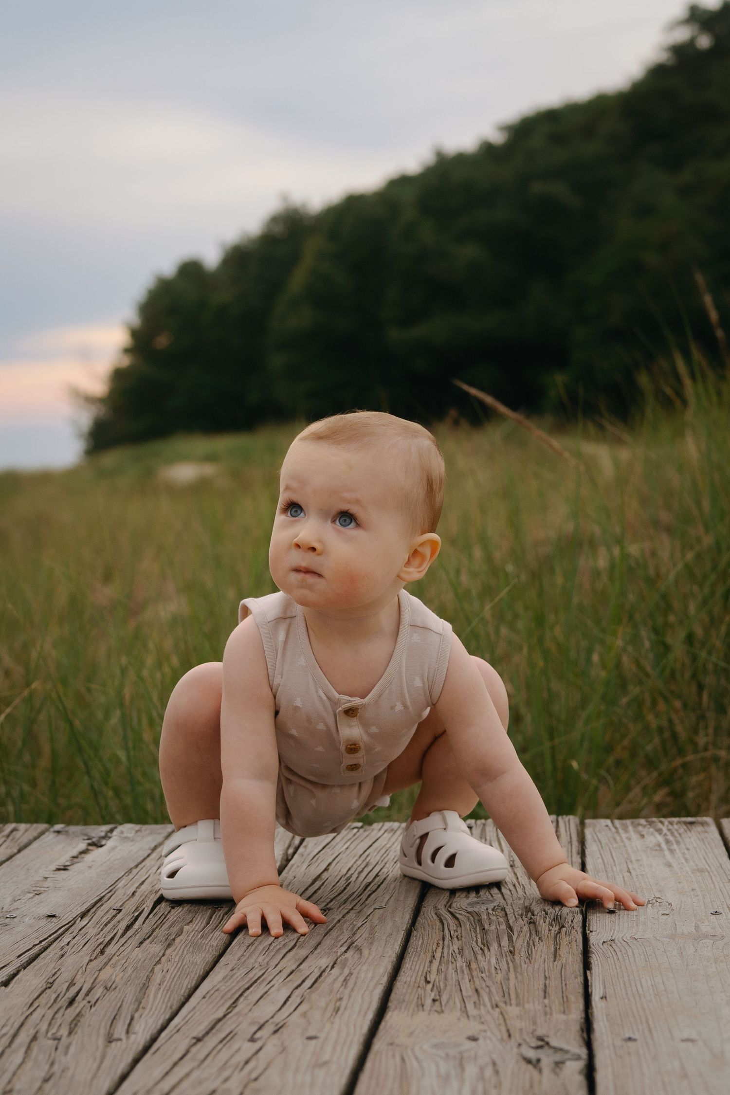 Baby in cream-colored outfit crawls on weathered wooden surface outdoors.