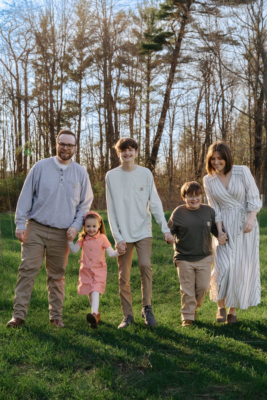 Happy family of five holding hands and walking together on a grassy field at sunset.
