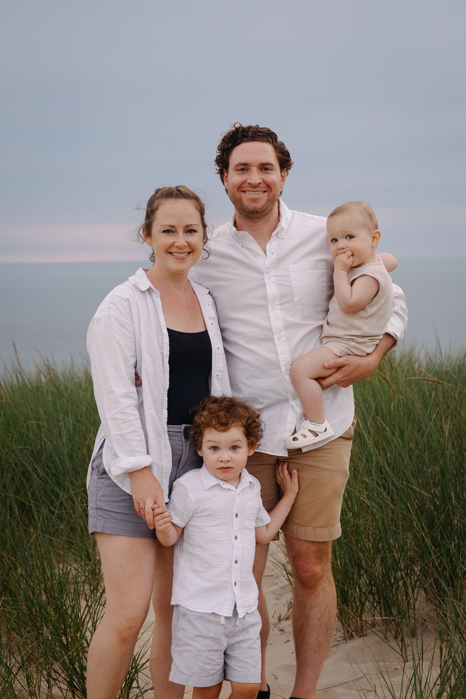 Family poses together on beach dunes wearing coordinated white and neutral outfits.