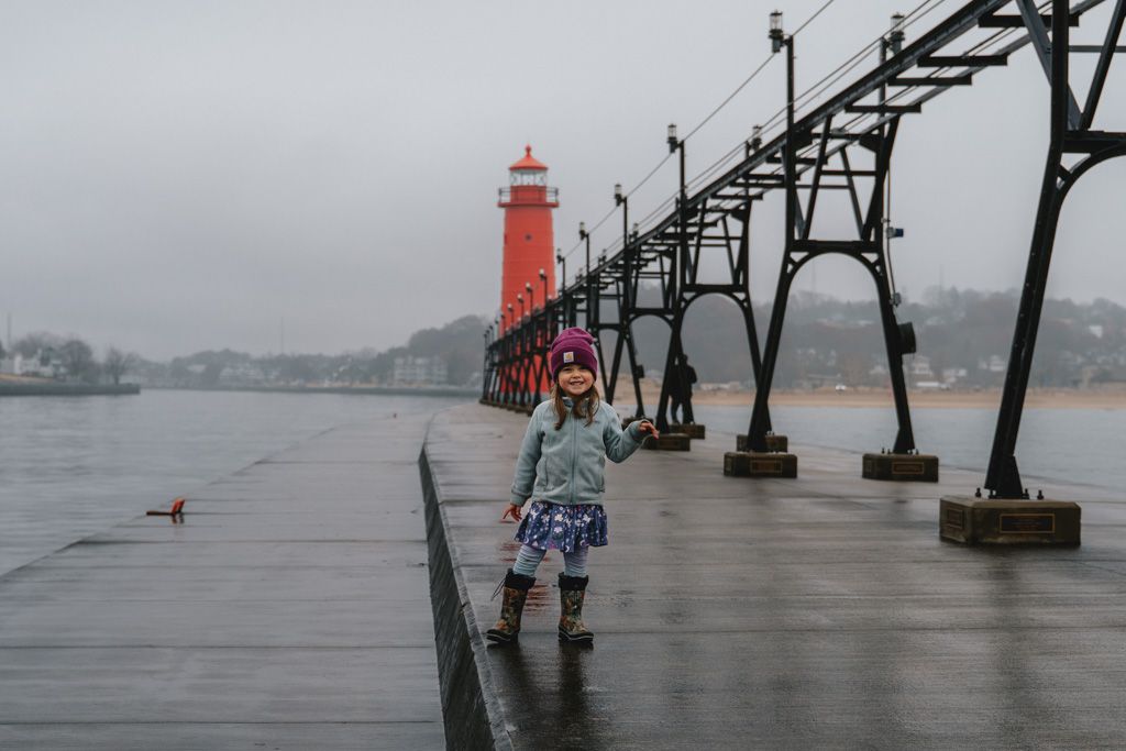 A young girl stands on the pier in front of a red lighthouse during an overcast day, creating a moody atmosphere.