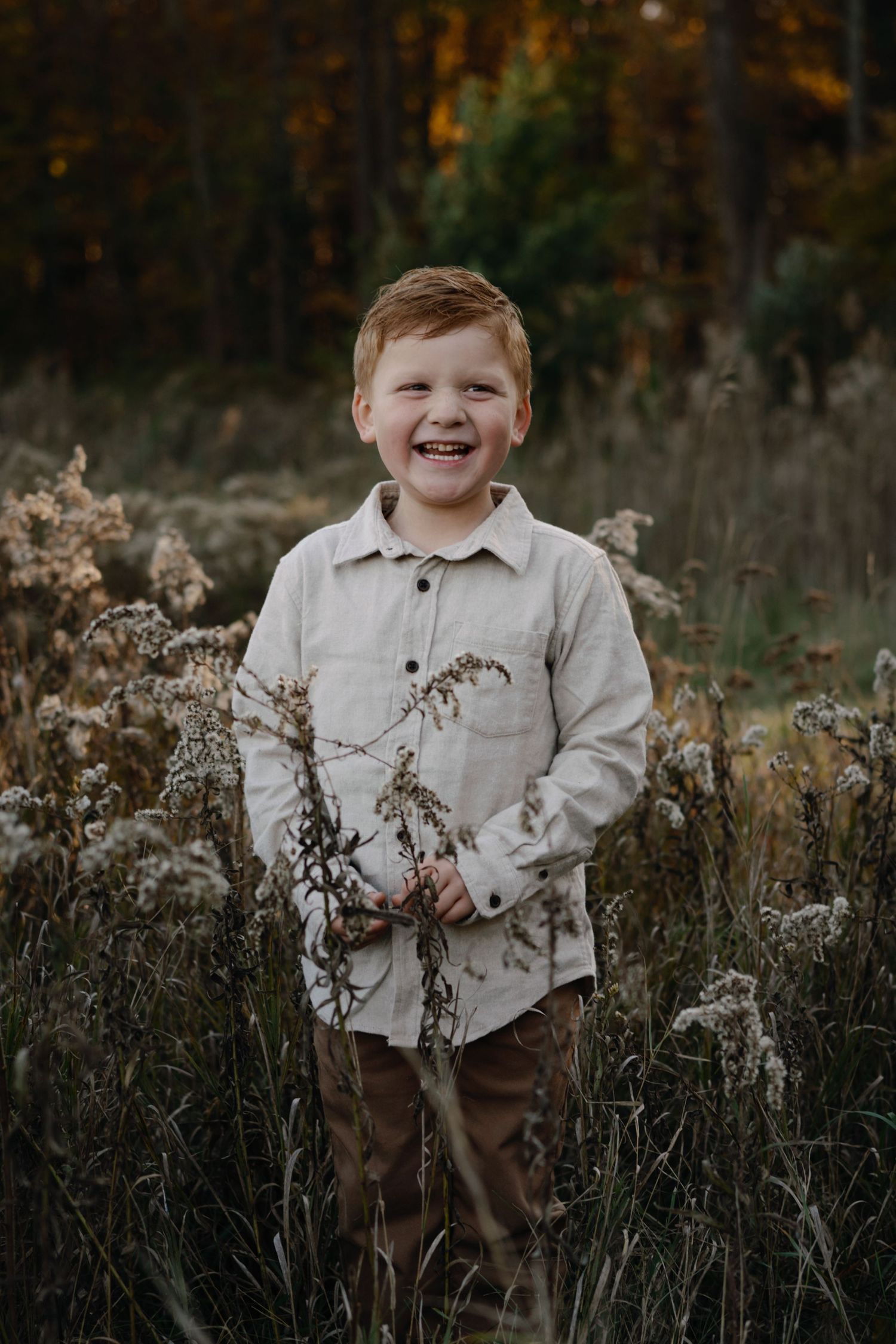 A child smiles while standing in a field of dried autumn plants wearing a light colored shirt.