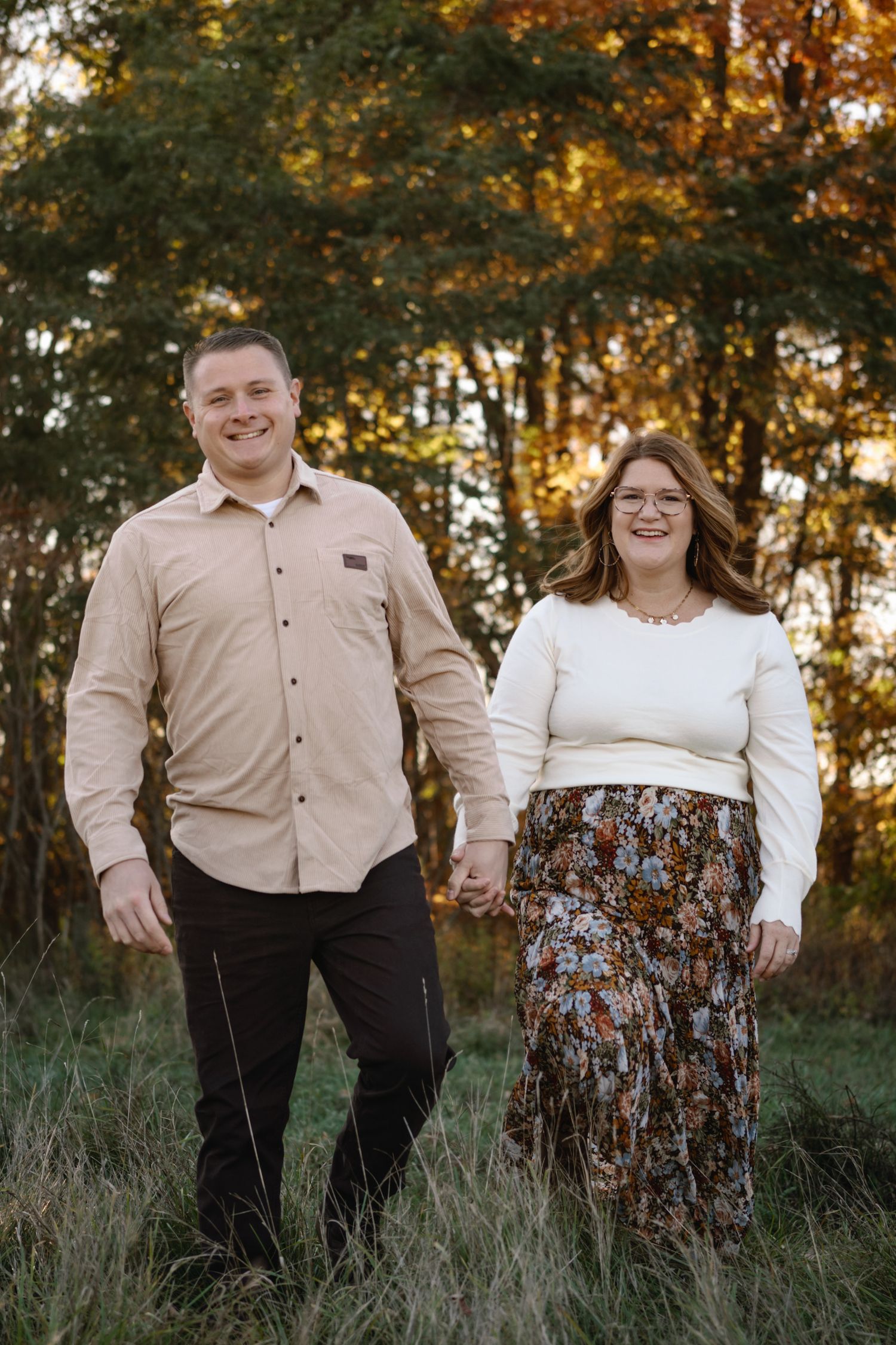 A couple stands together in a field during golden hour wearing coordinated beige and brown outfits.