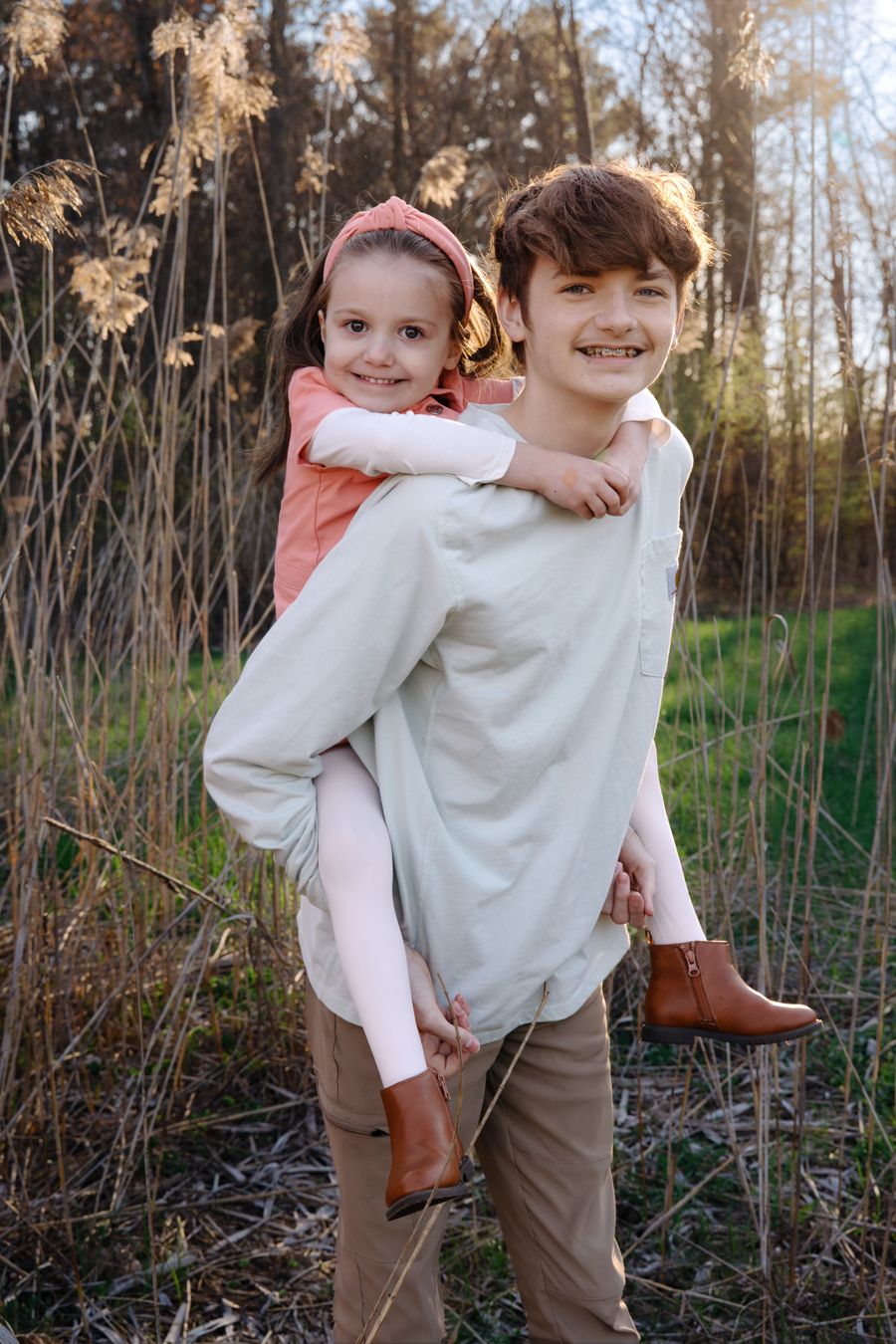 A smiling boy gives a young girl a piggyback ride outdoors among tall dry grasses and trees.