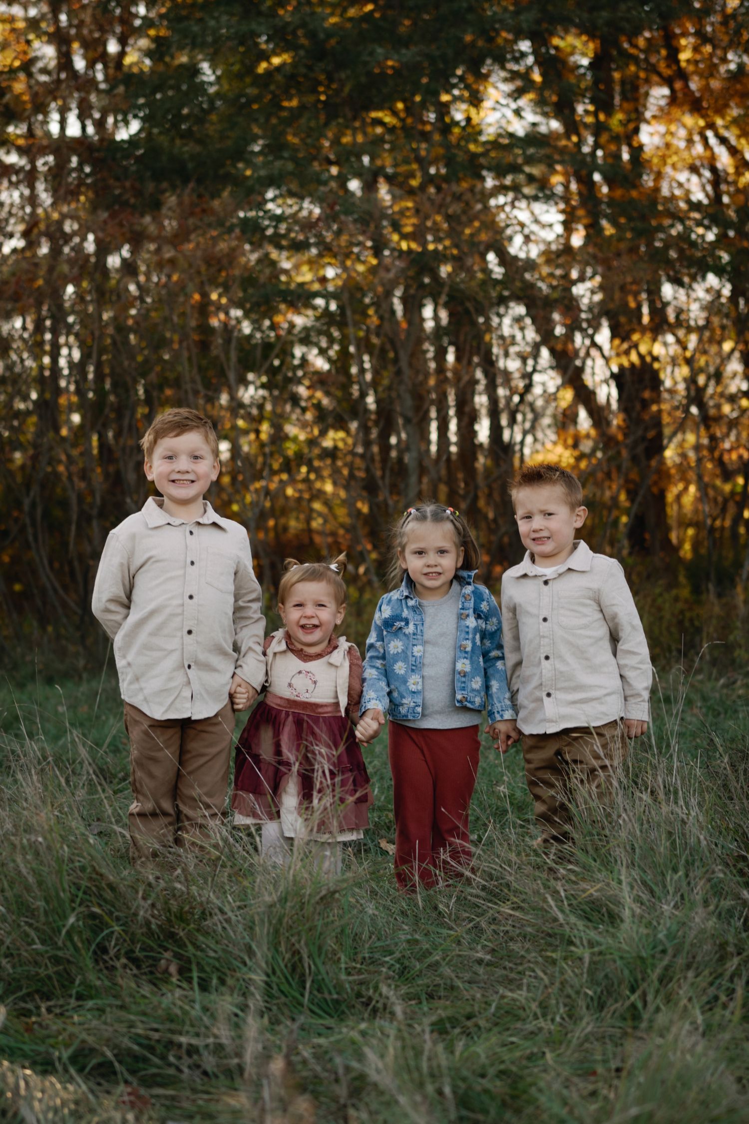 Four young children holding hands in a grassy field with autumn trees, wearing coordinated fall outfits.