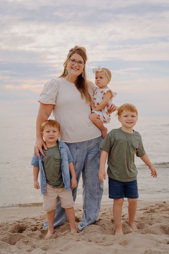 A family poses together on a sandy beach at sunset wearing casual clothing in coordinating neutral colors.