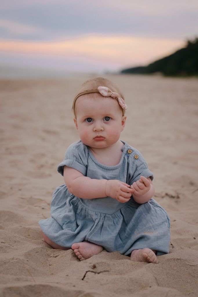 Baby in light blue dress sits on sandy beach during sunset creating a peaceful coastal portrait.