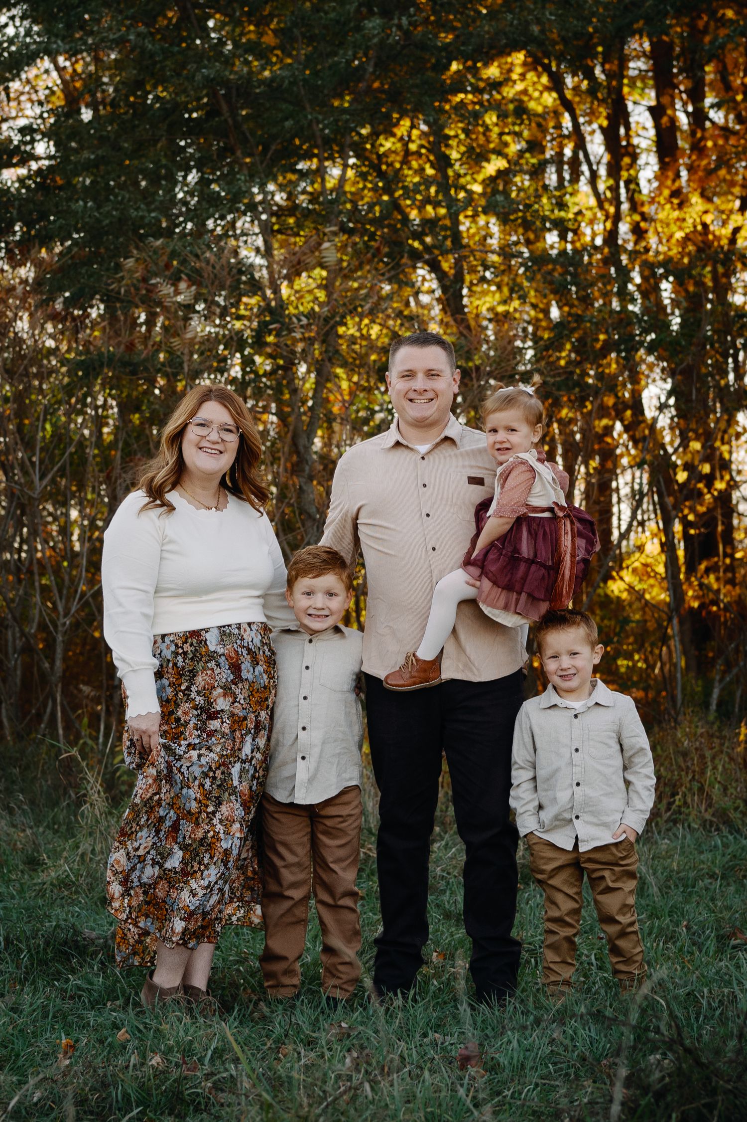 Family group photo with coordinated outfits standing in tall grass during golden hour.