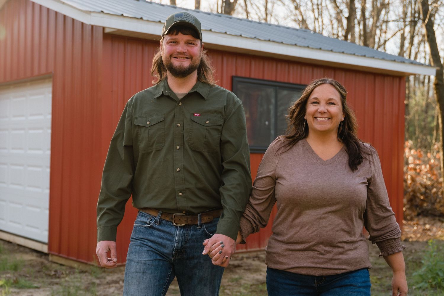 A smiling couple holds hands outdoors in front of a red barn building surrounded by trees.