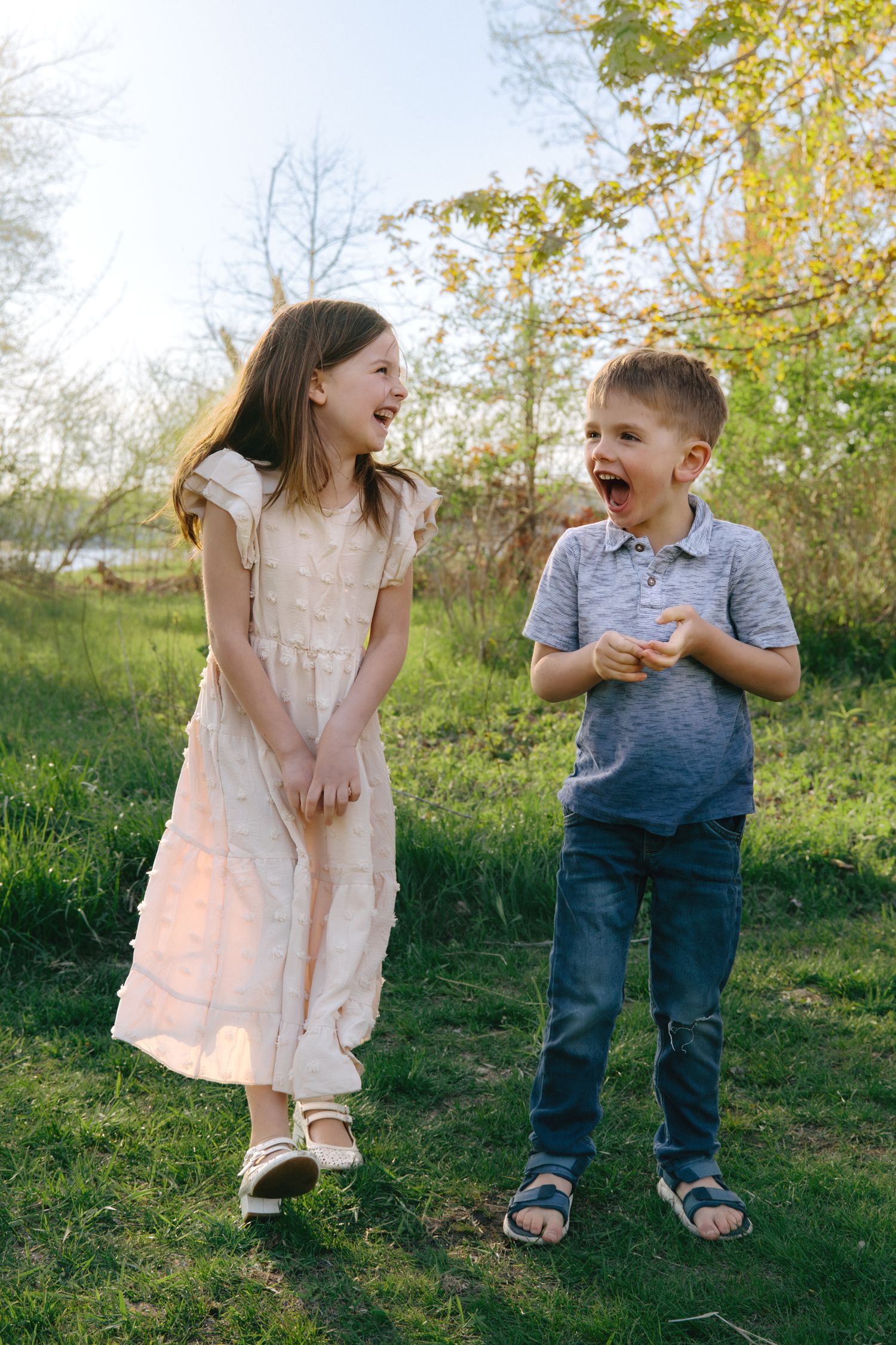 A girl in a white dress and a boy in a gray shirt laugh together while standing on green grass outdoors.