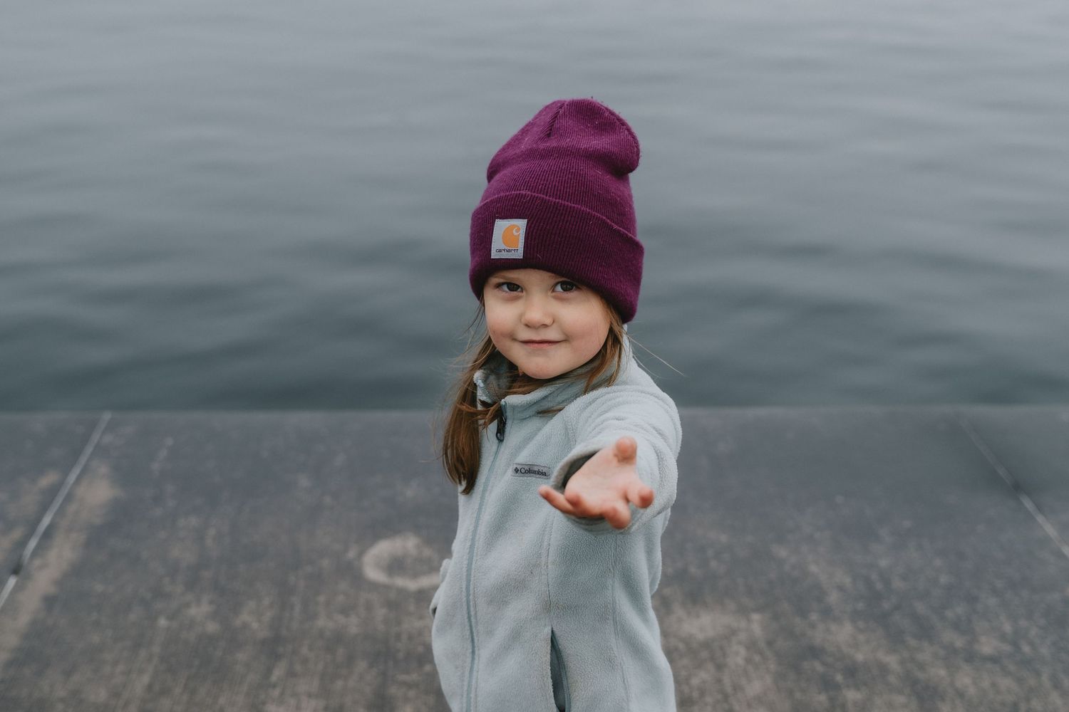 A child wearing a burgundy beanie and gray sweatshirt stands by a lake on an overcast day.