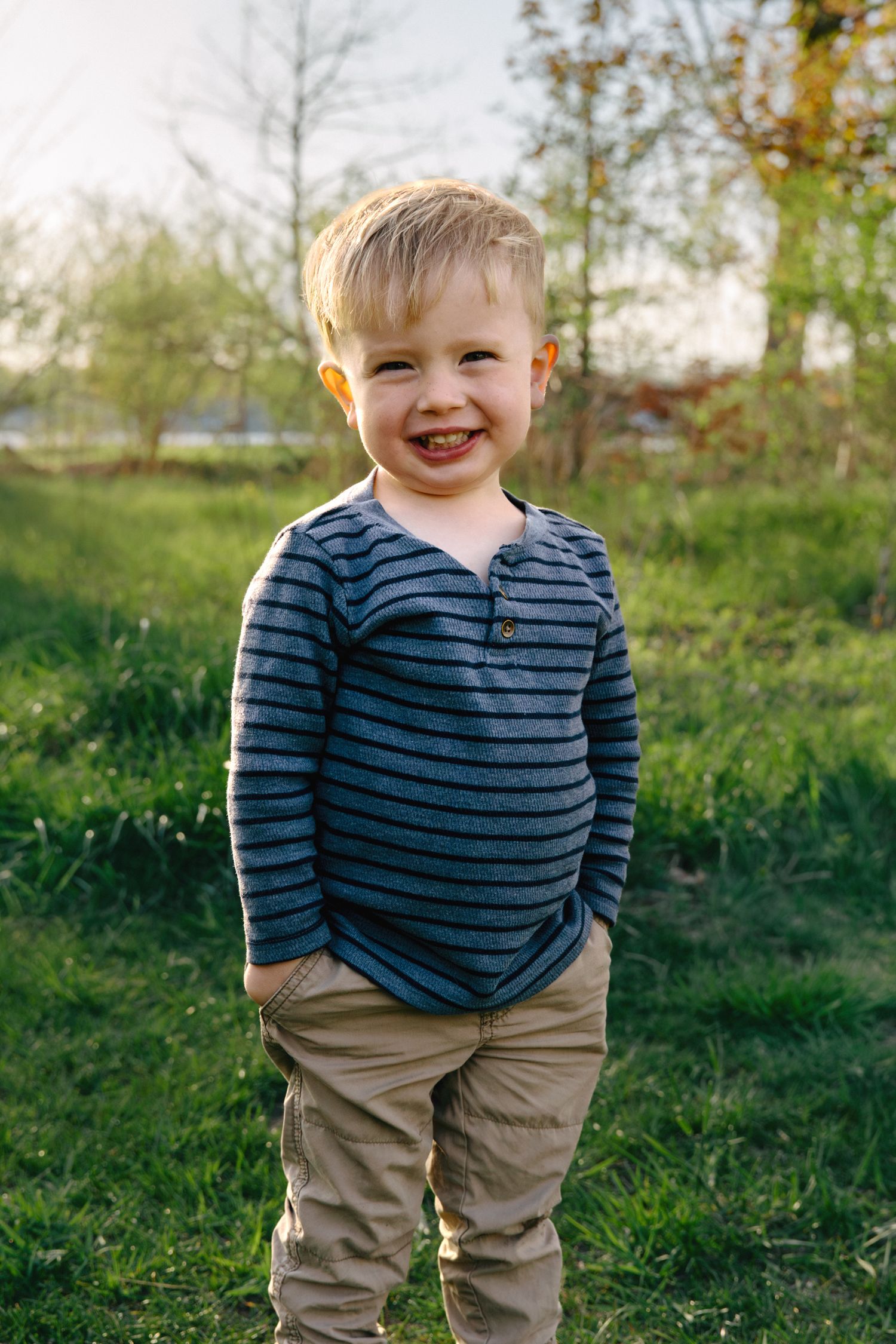A cheerful toddler boy in a striped shirt and khaki pants smiles outdoors in a lush green grassy field.