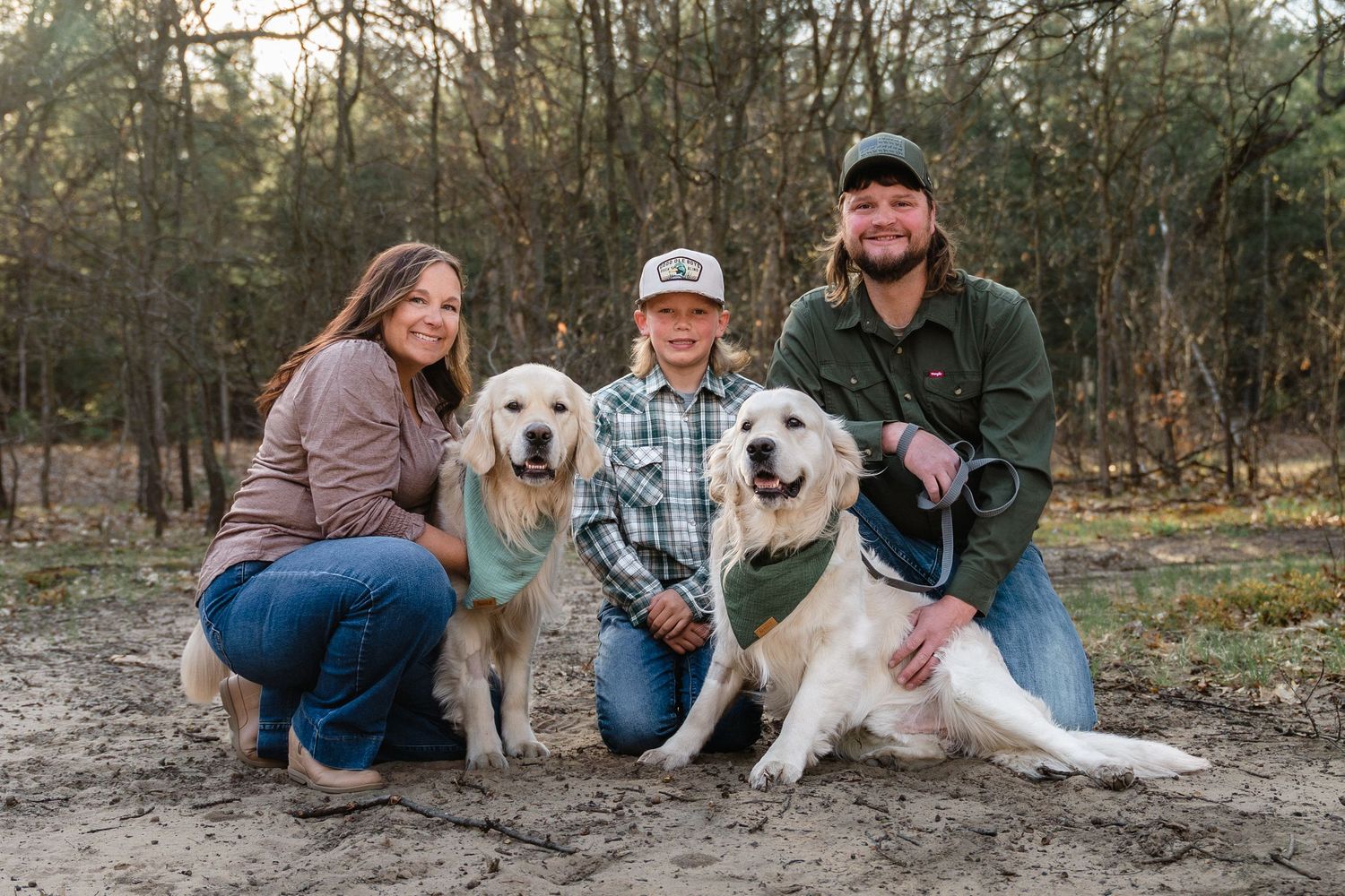 Family of three poses with two golden retrievers outdoors in a wooded sandy area during golden hour.