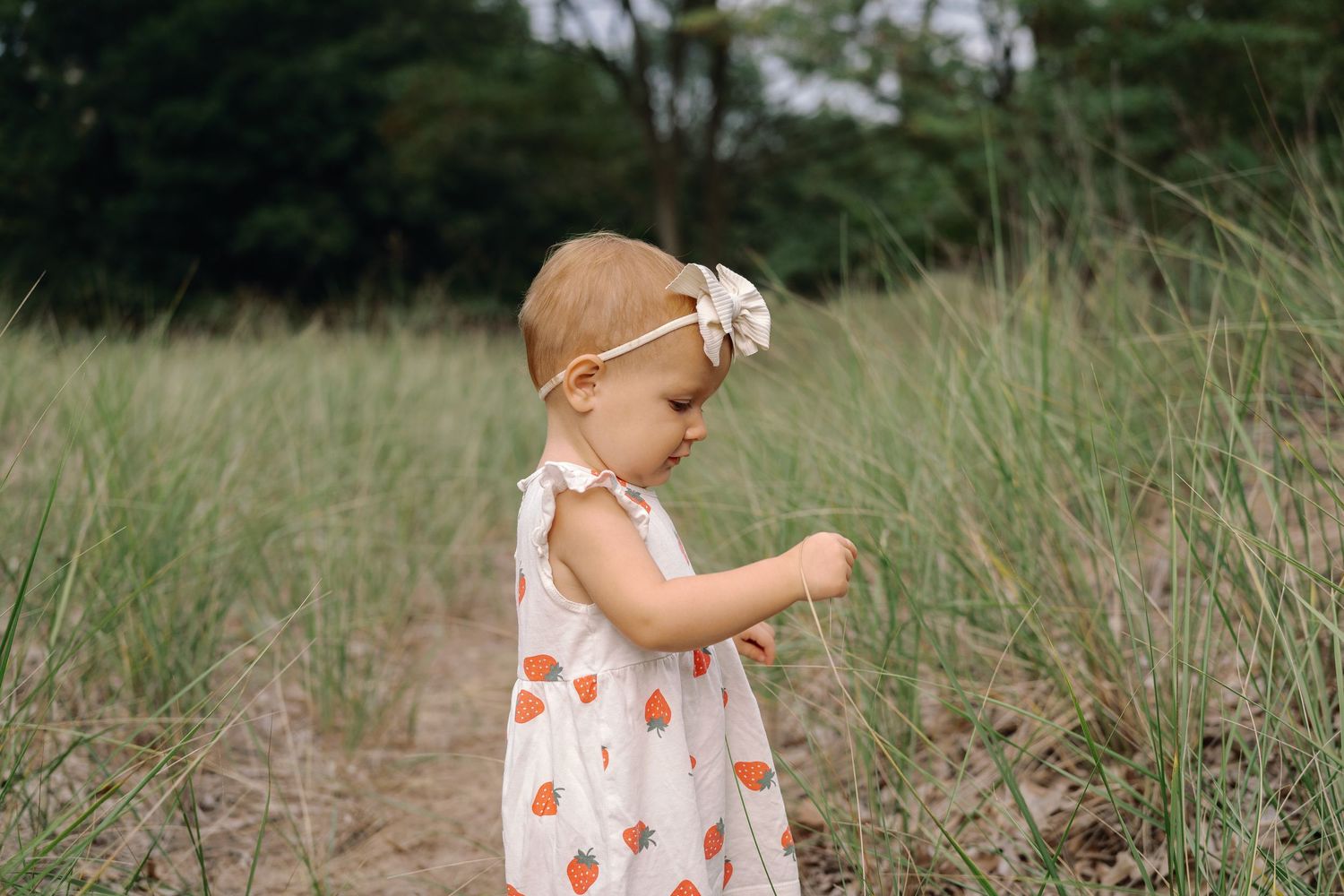 A small child explores nature wearing a white dress with orange hearts amid green grass and trees.