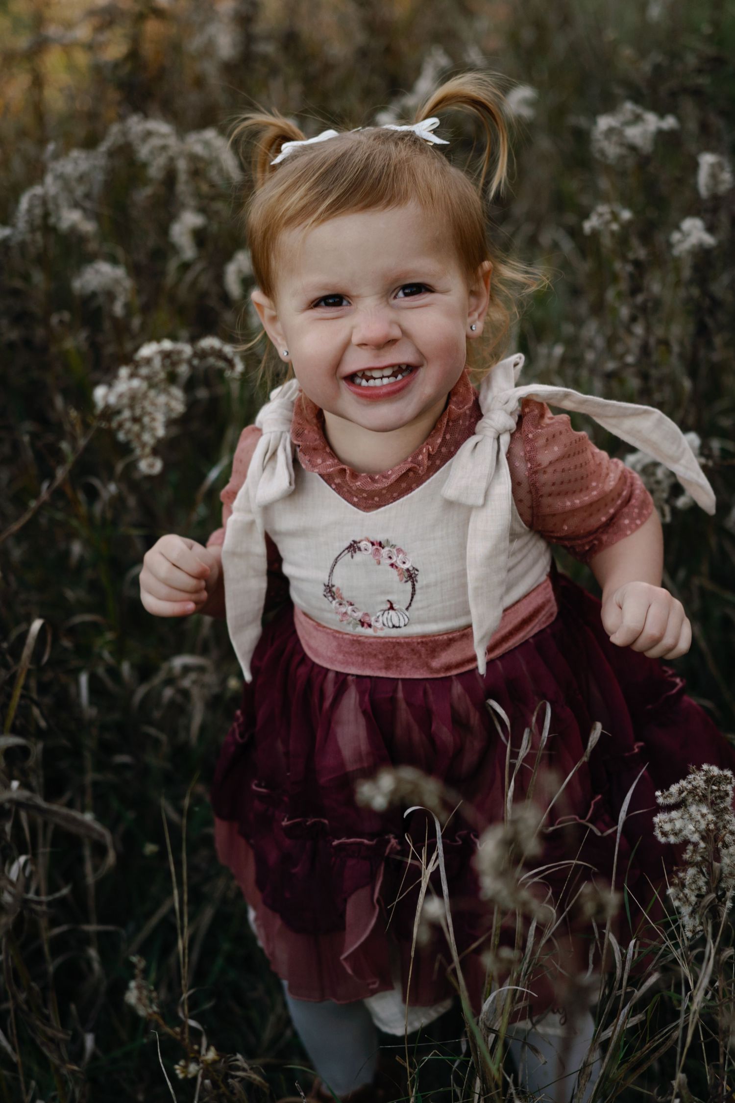 Child wearing burgundy dress and white collar explores tall grasses and wildflowers.