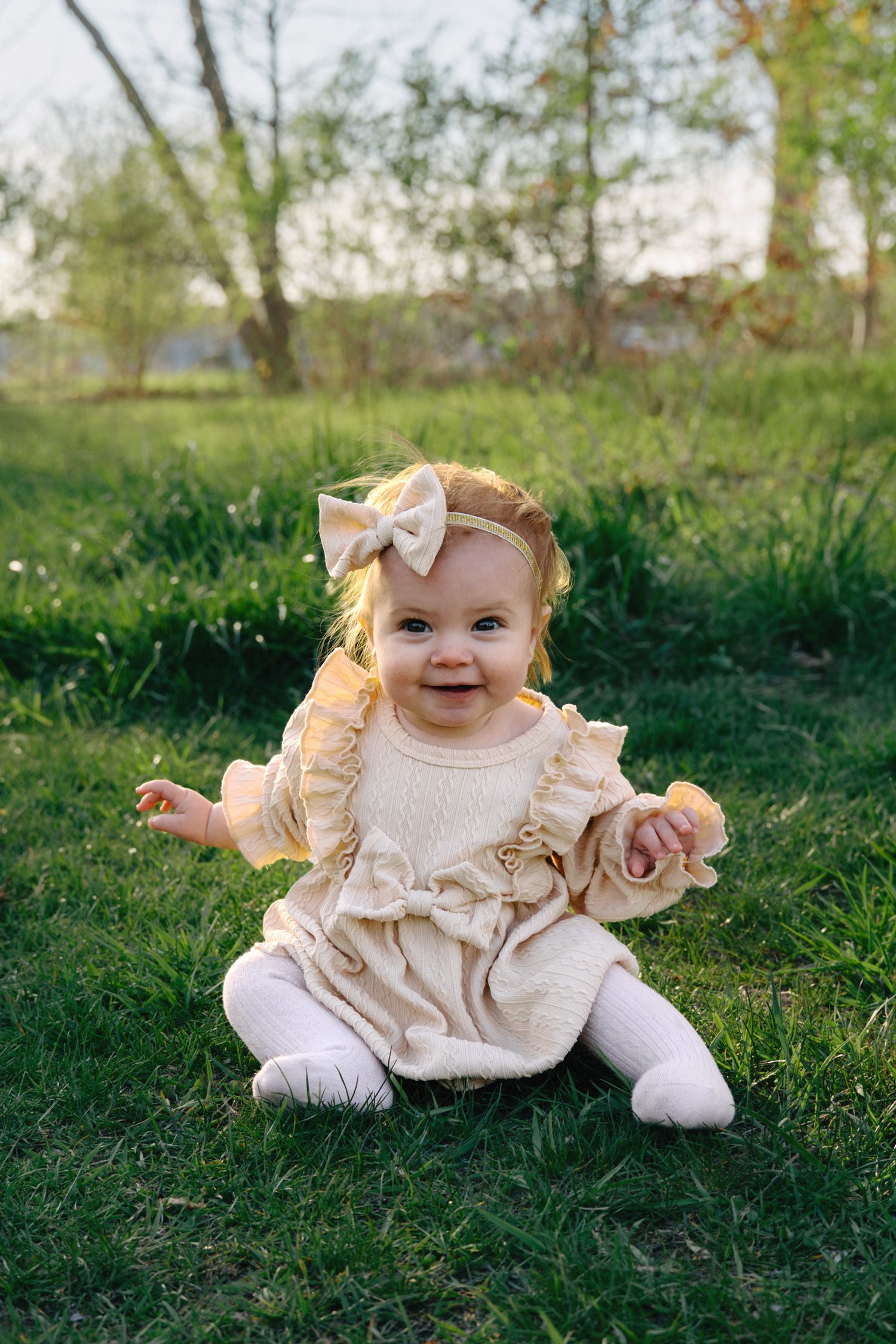 Smiling baby girl in cream ruffle dress and bow headband sitting on green grass outdoors in warm sunlight.