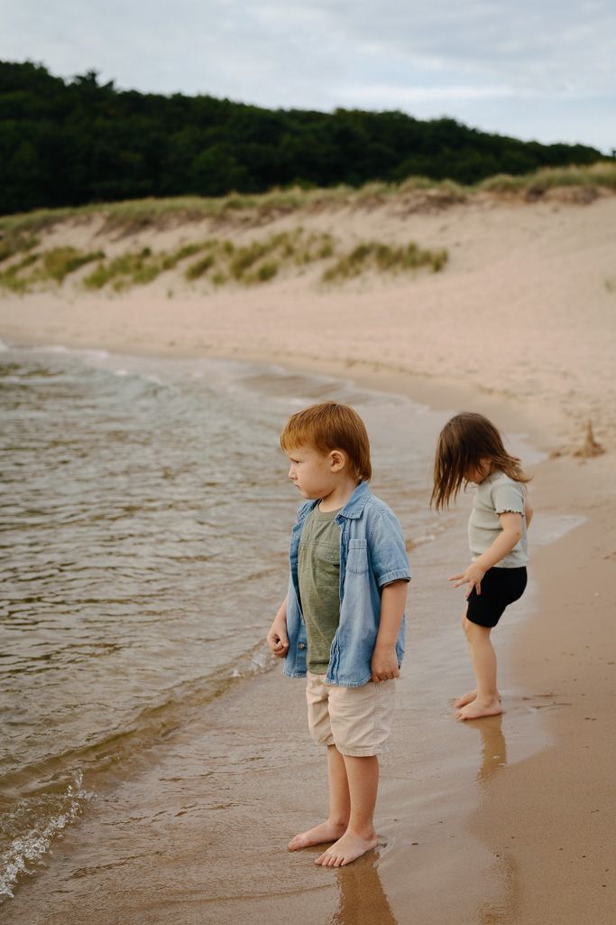 Two young children play at the water's edge on a sandy beach with dunes in the background during golden hour.