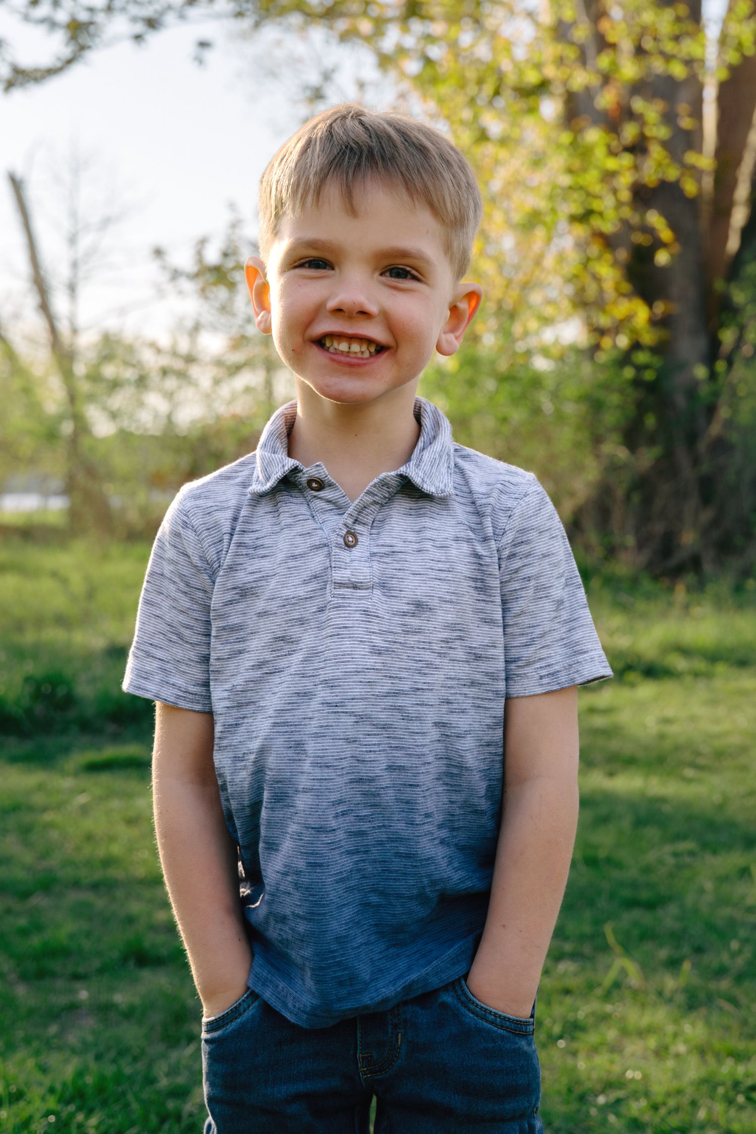 Smiling young boy with blonde hair wearing a gray polo shirt and jeans standing outdoors in a green yard.