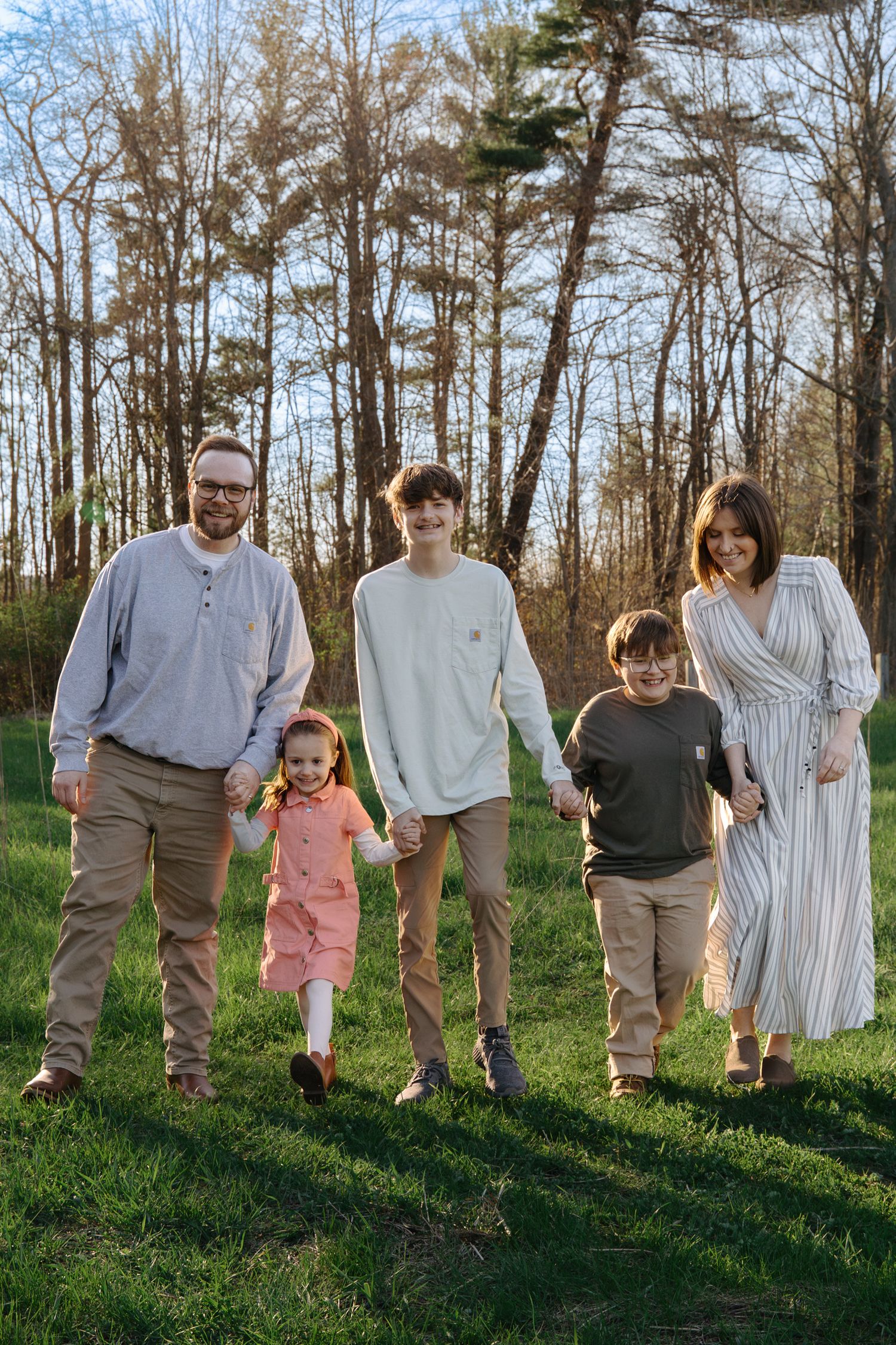 Family of five holding hands walking on green grass with trees in background on a sunny day.