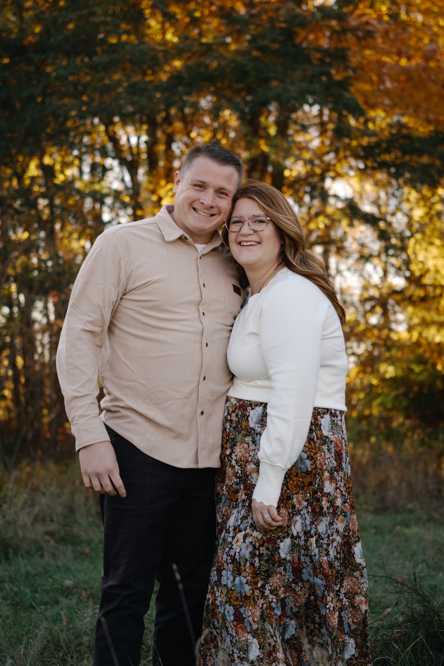 A couple poses together in an autumn setting with golden foliage in the background.