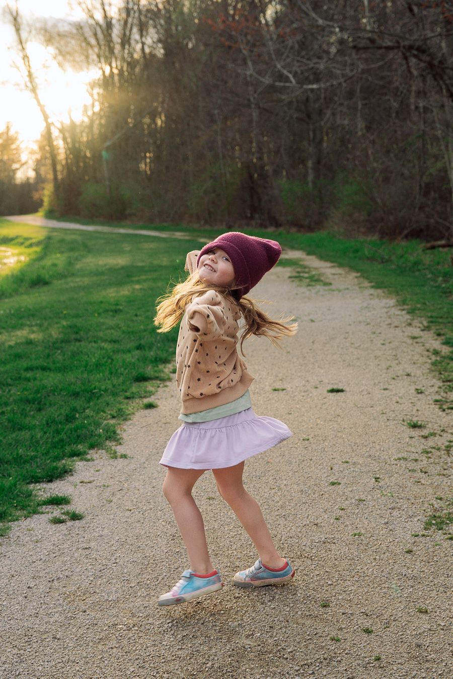 A joyful young girl in a beanie and polka dot jacket twirls on a sunlit park path surrounded by green grass.