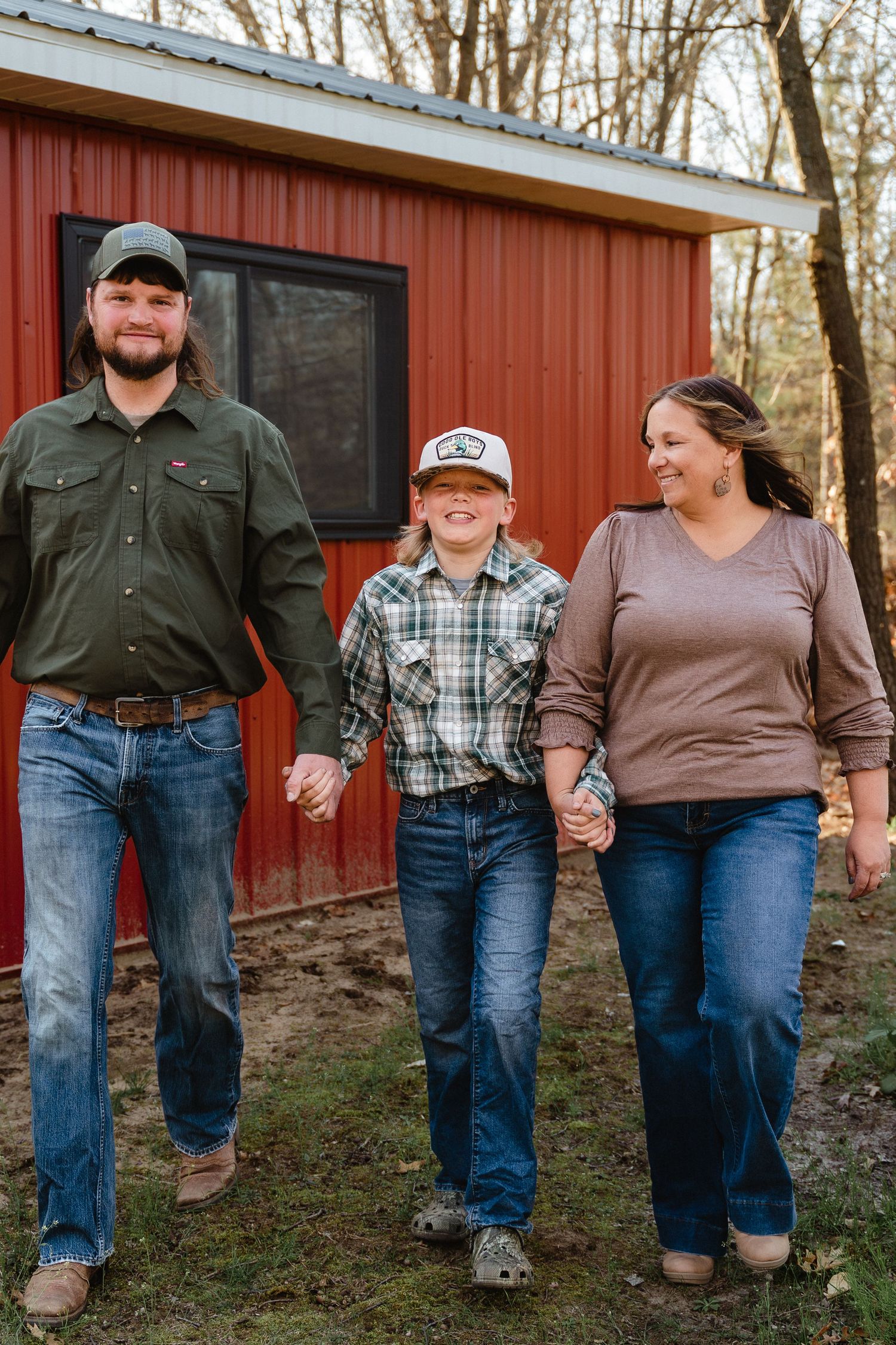 A happy family of three holding hands, walking in front of a red barn, wearing casual country attire.