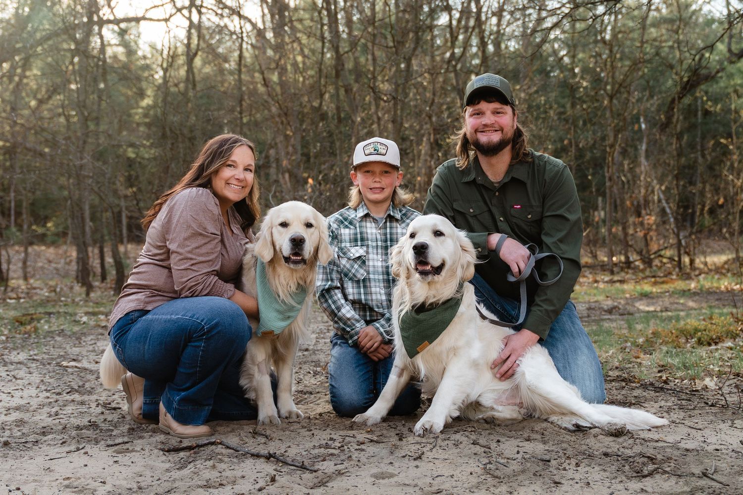 Family of three posing outdoors with two golden retrievers in a wooded sandy area.