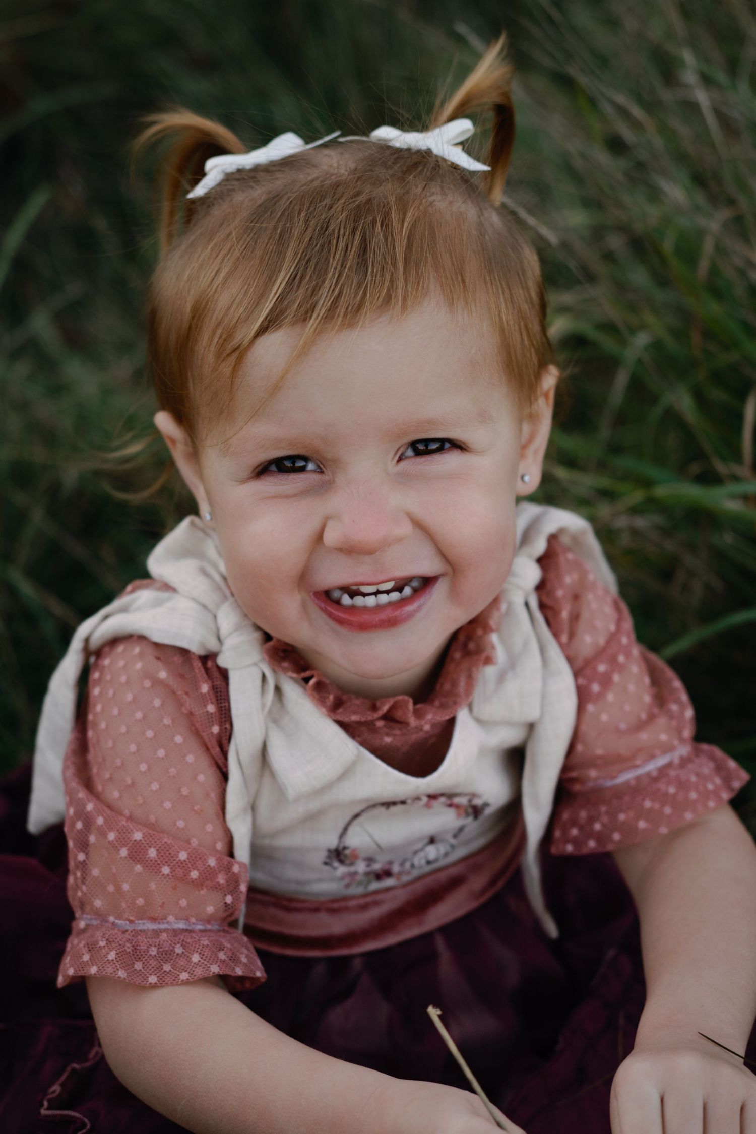 A young child in a pink dress smiles joyfully while sitting in grass.