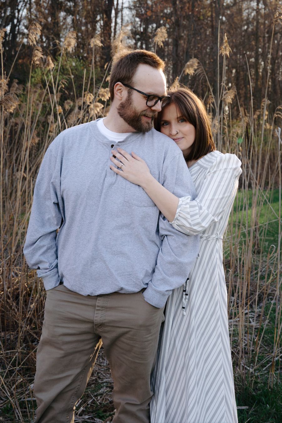 A couple embraces tenderly outdoors, the woman in a striped dress leaning on a bearded man in a gray sweatshirt.