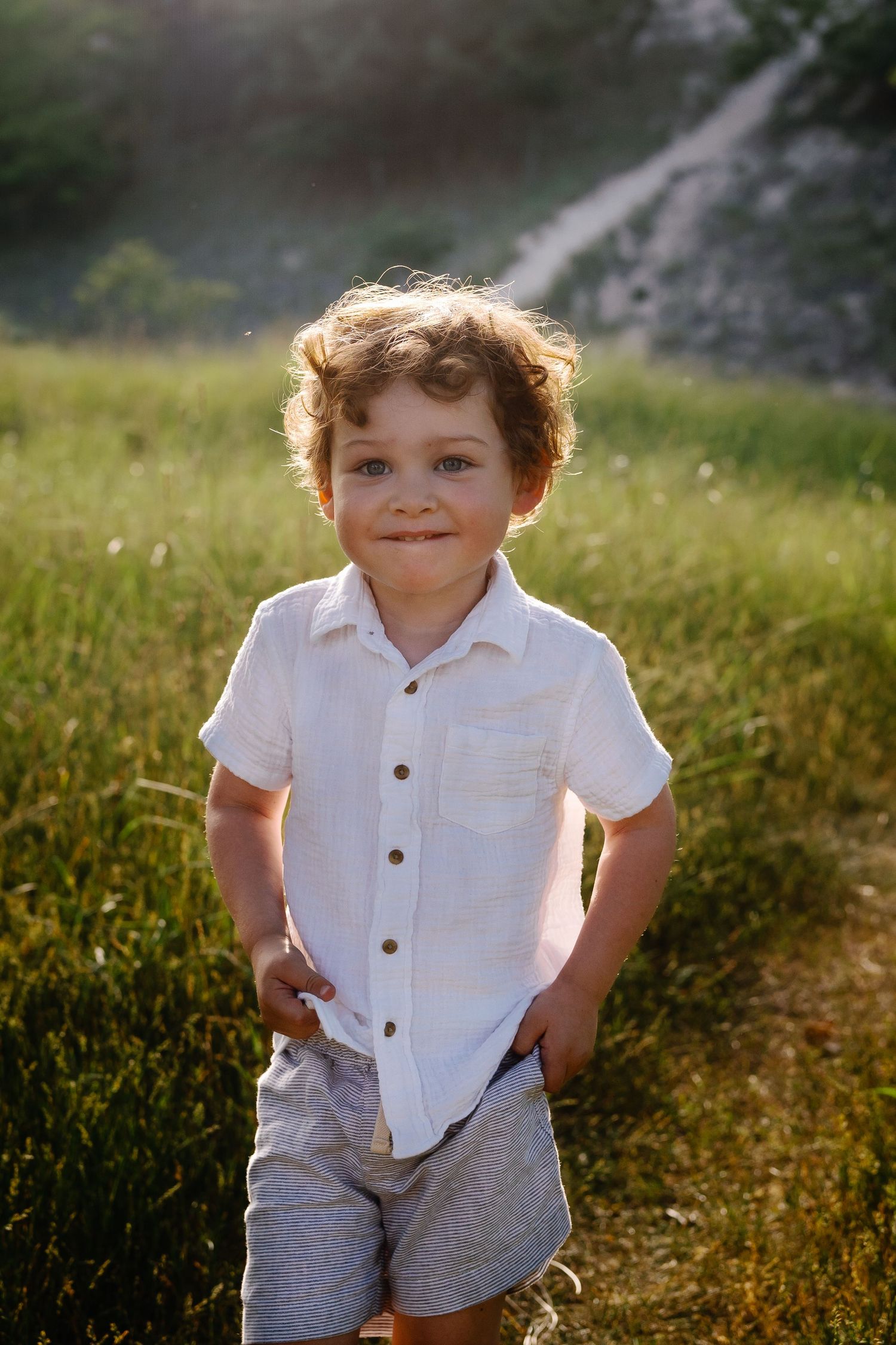 Child in white button-up shirt and grey shorts plays in sunny summer field.