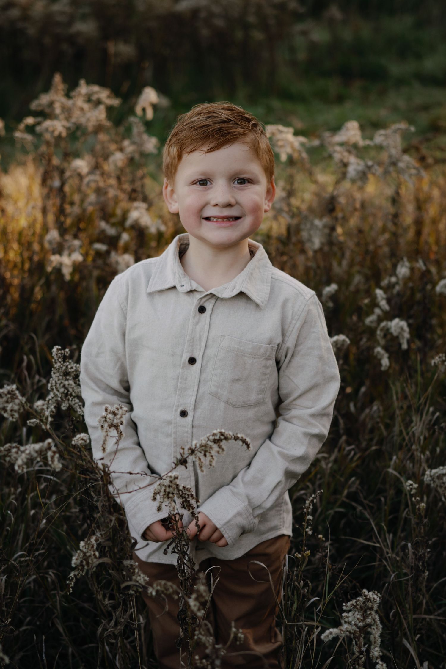 A child in a beige button-down shirt poses among dried wildflowers during golden hour.