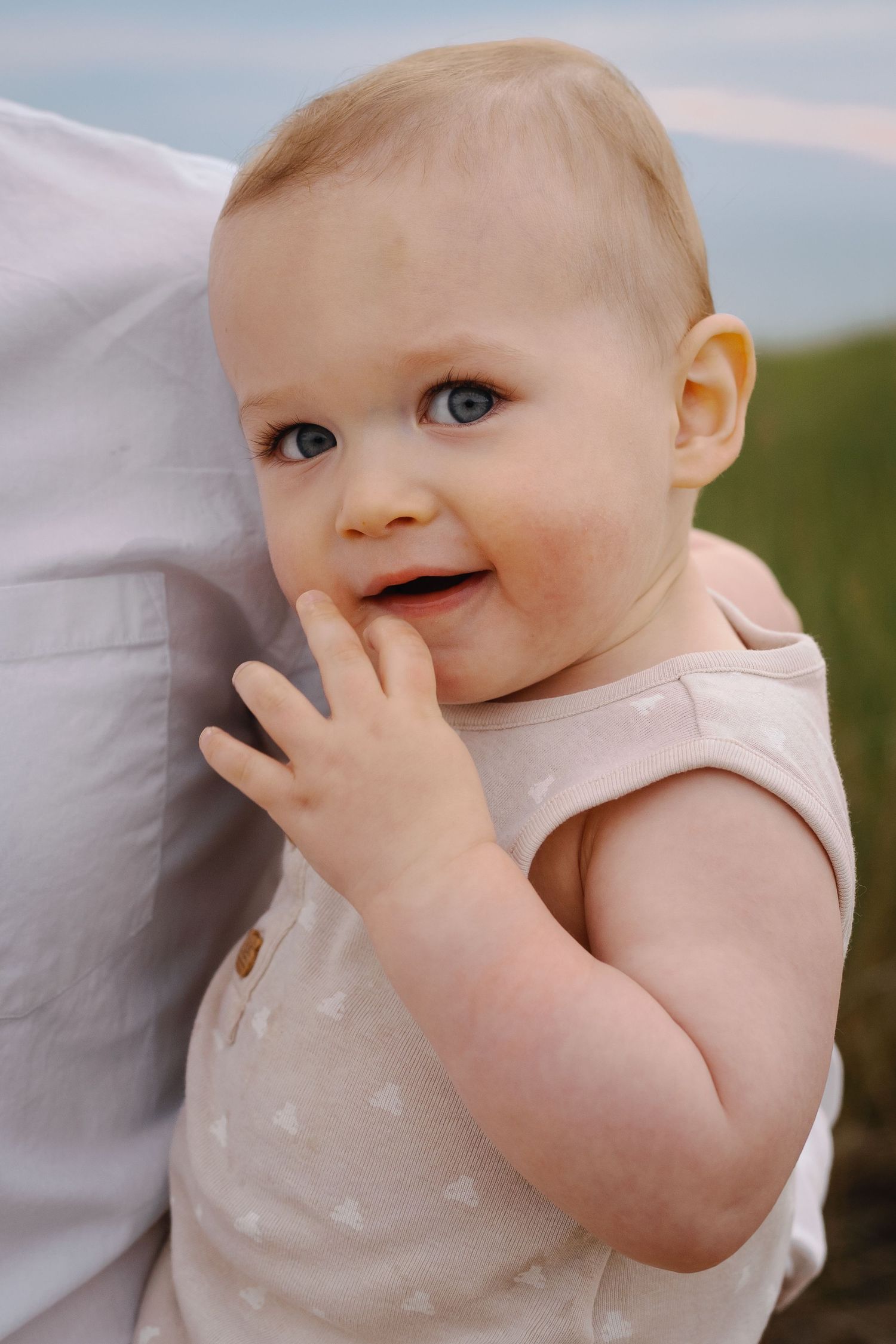 Baby in light-colored outfit smiles while touching their face, captured in close-up.