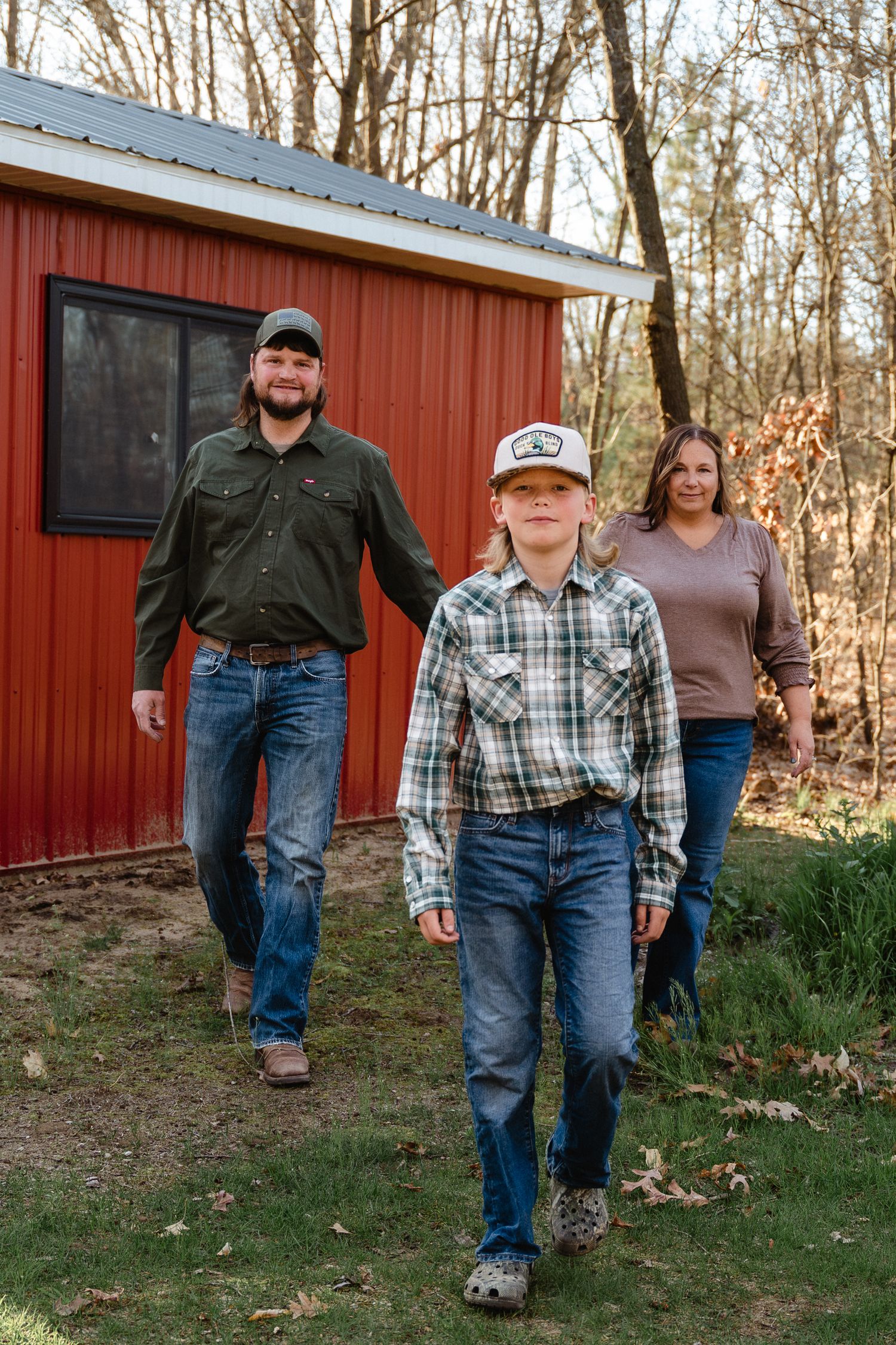 A family of three walks toward the camera in front of a red barn in a wooded yard.