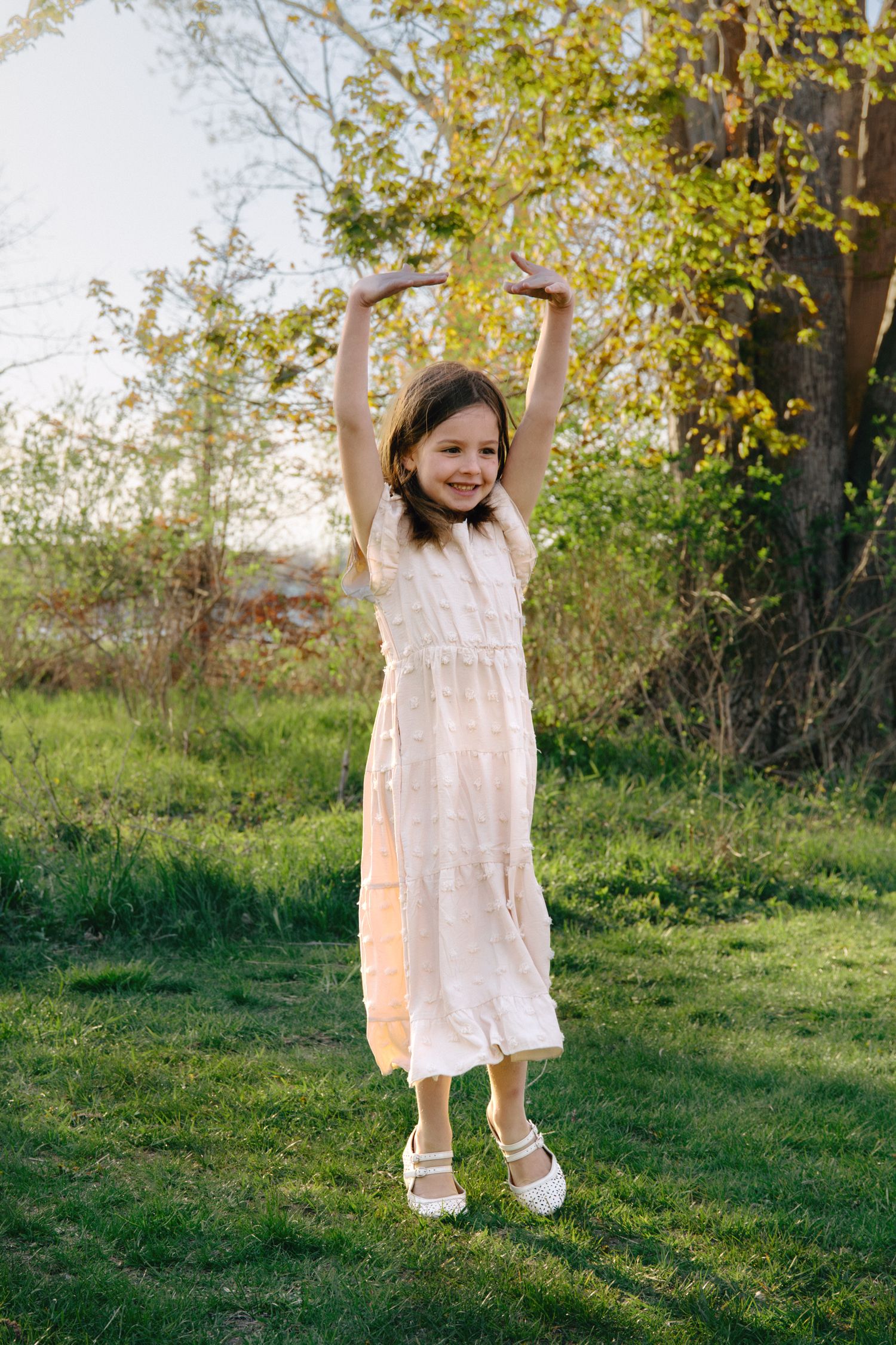 Young girl in white dress dancing outdoors on green grass with arms raised, bathed in golden sunlight.