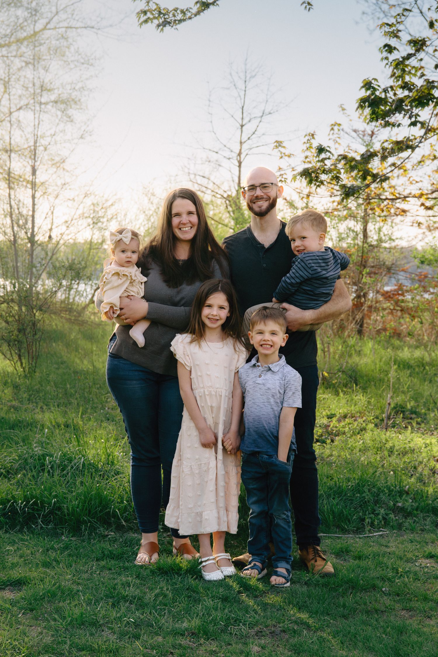Family of six posing outdoors on green grass with trees and soft sunset light in background.