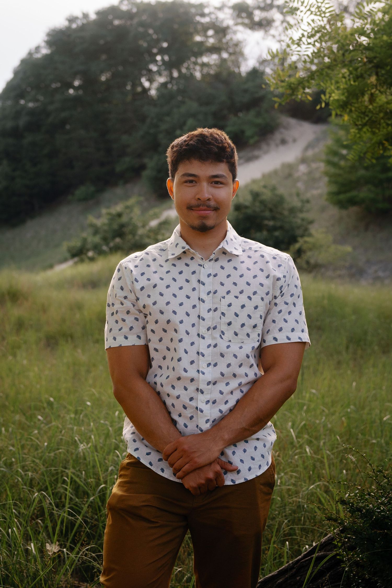 Young man in a patterned white shirt and brown pants poses casually in a grassy field at sunset.