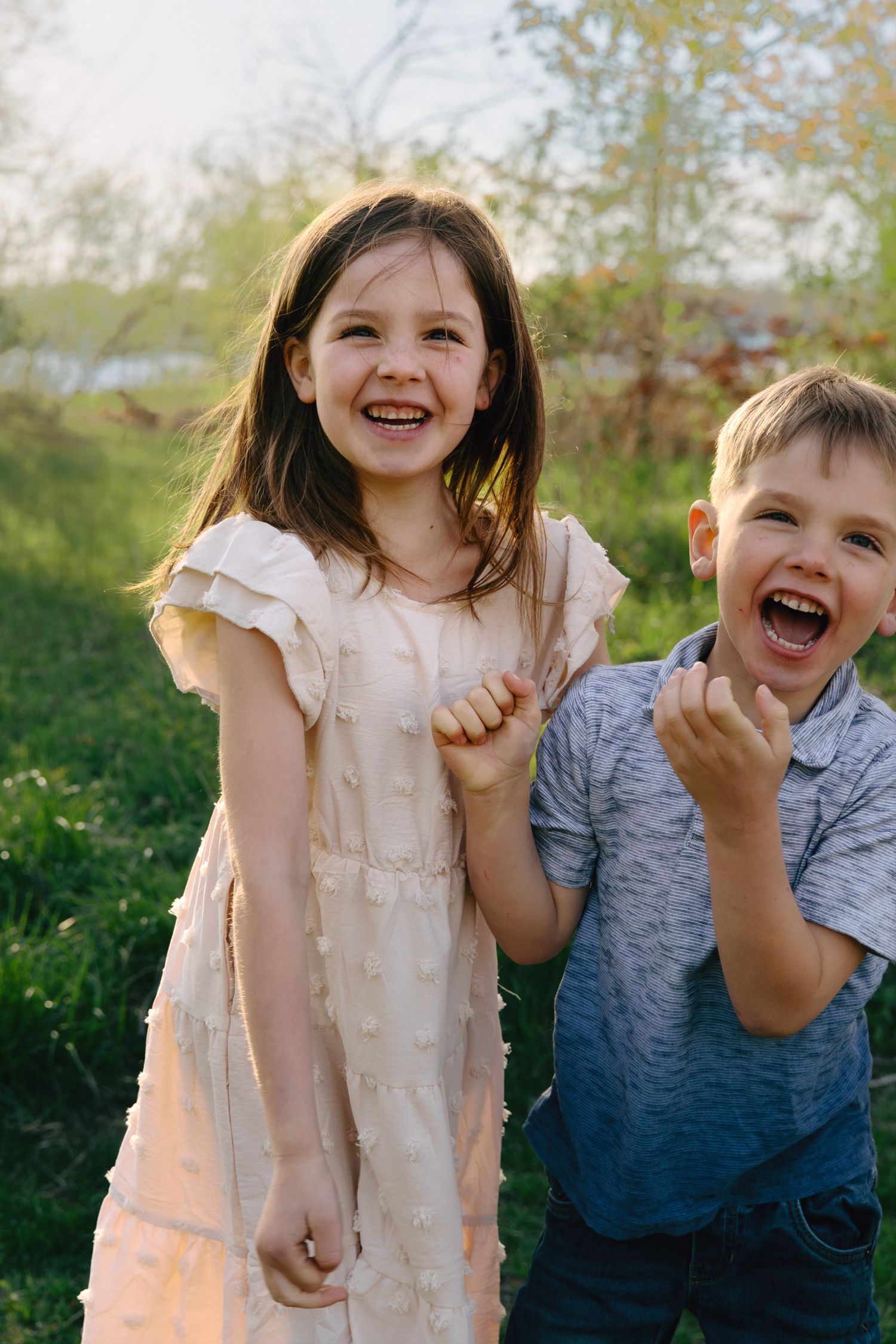 Two laughing children, a girl in a white dress and a boy in a gray shirt, play outdoors in a sunny park.