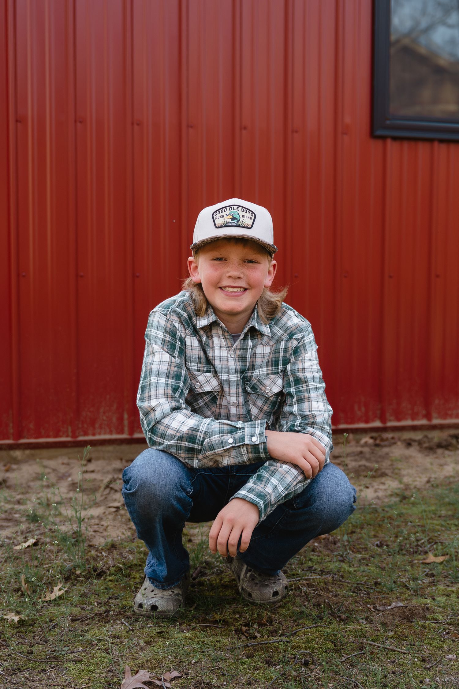 Smiling boy in plaid shirt and trucker hat crouching in front of a red barn wall outdoors.