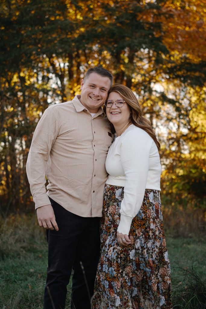 Couple in autumn setting with fall foliage backdrop wearing coordinated beige and white outfits.