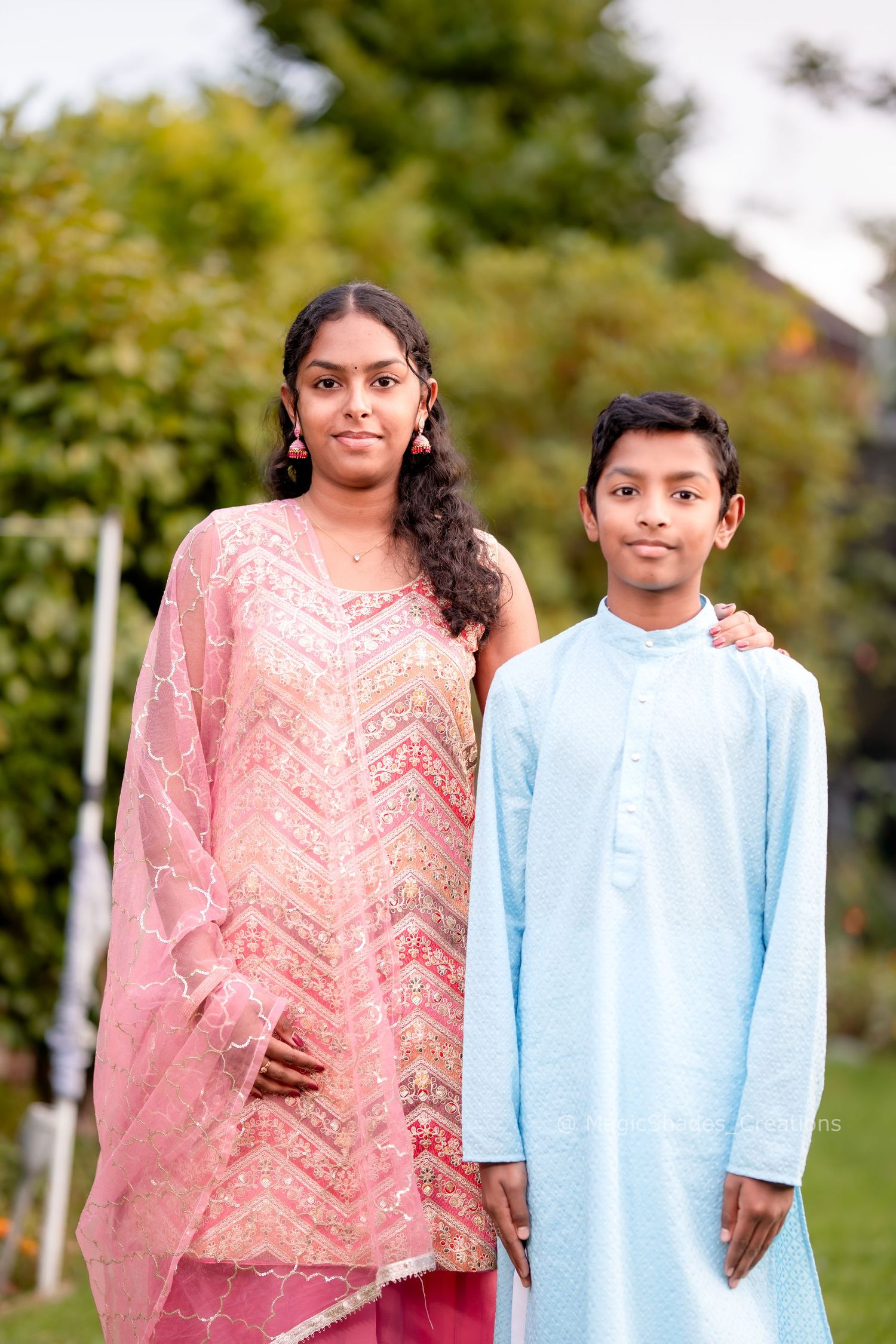 Two children in traditional South Asian attire pose outdoors, a girl in pink salwar and a boy in light blue kurta.