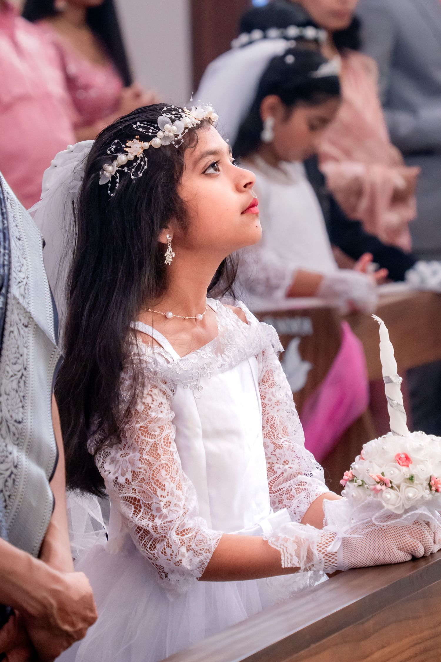 Young girl in white lace dress and floral crown holds bouquet during First Communion ceremony in church.