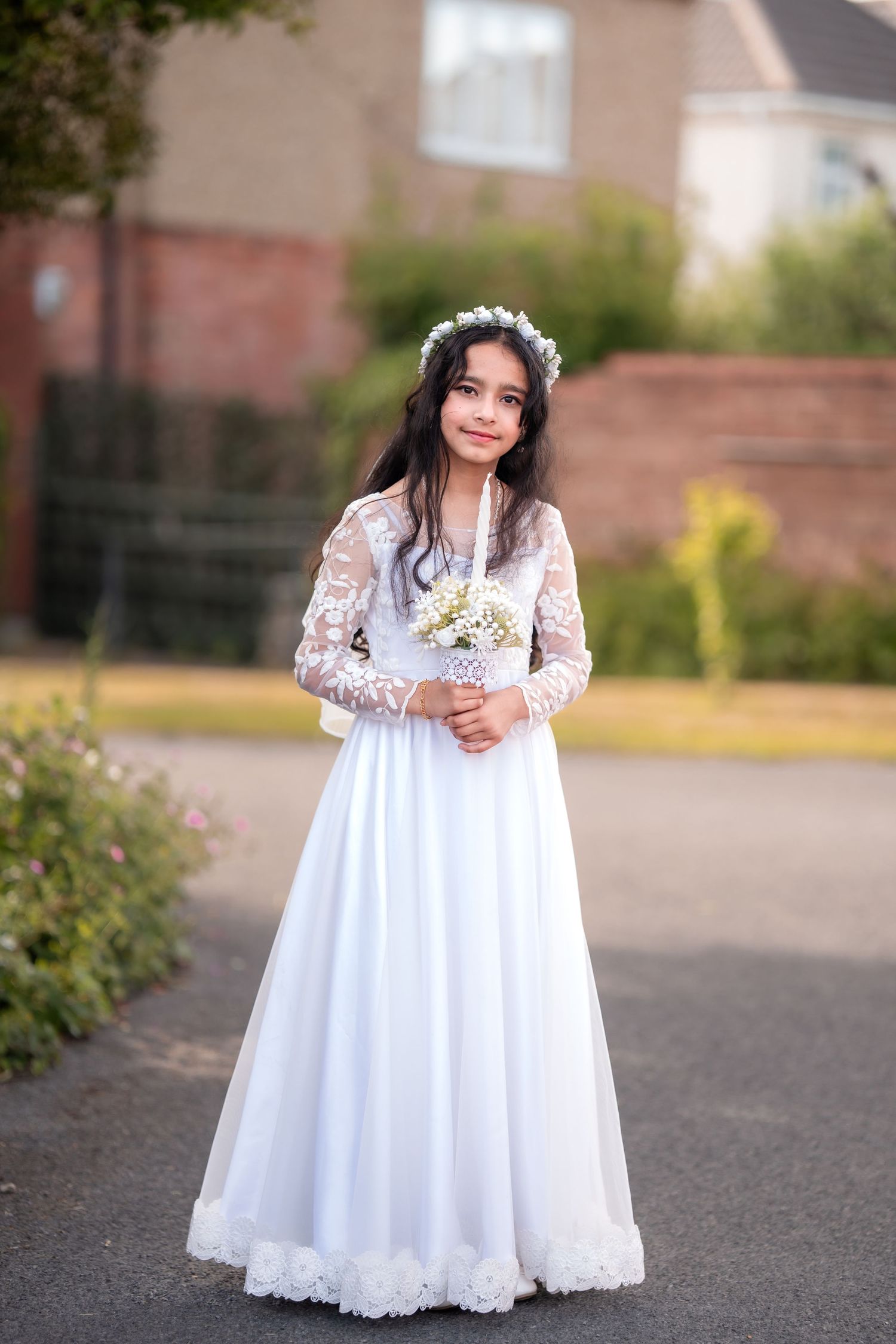 Young girl in white lace communion dress and floral crown holds bouquet outdoors.