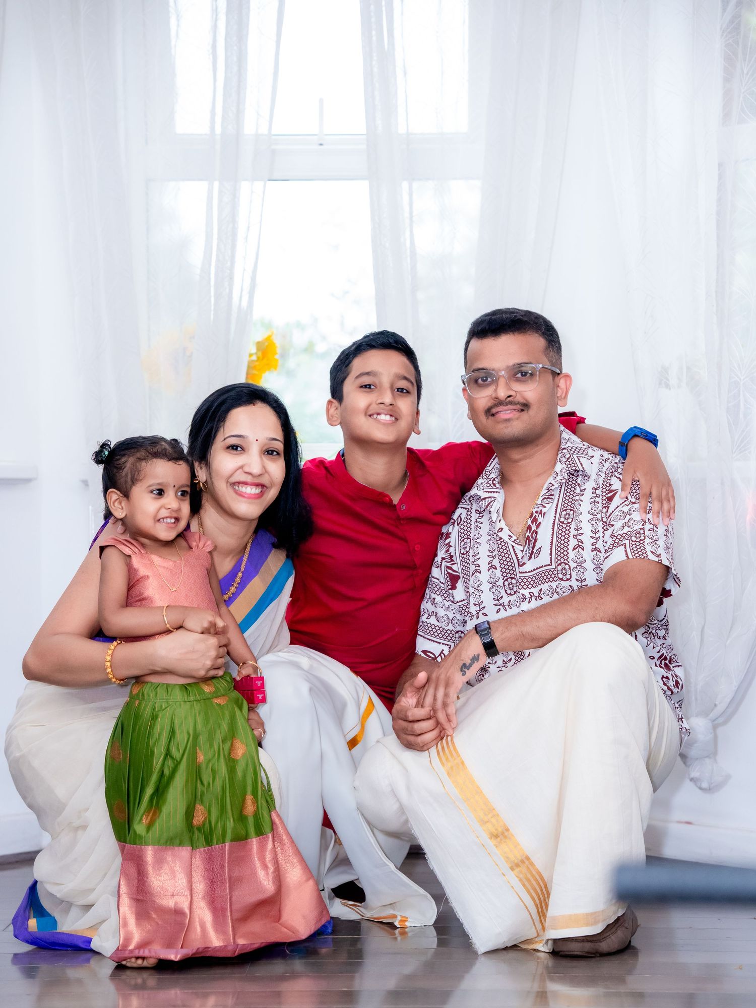 Indian family of four in traditional attire smiling together, parents with young daughter and son posing indoors.