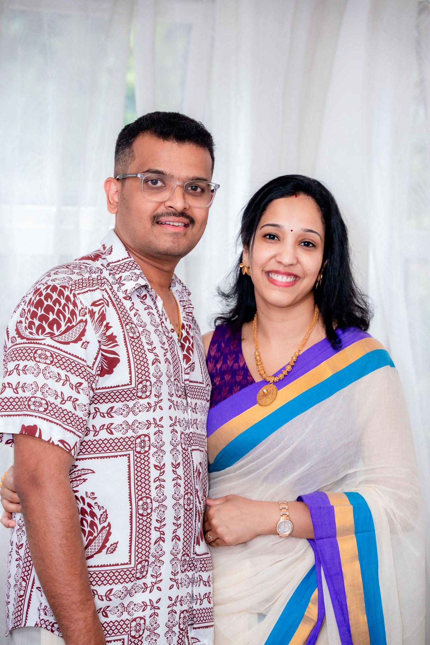 Indian couple smiling in traditional attire, man in printed shirt, woman in elegant white and purple saree.