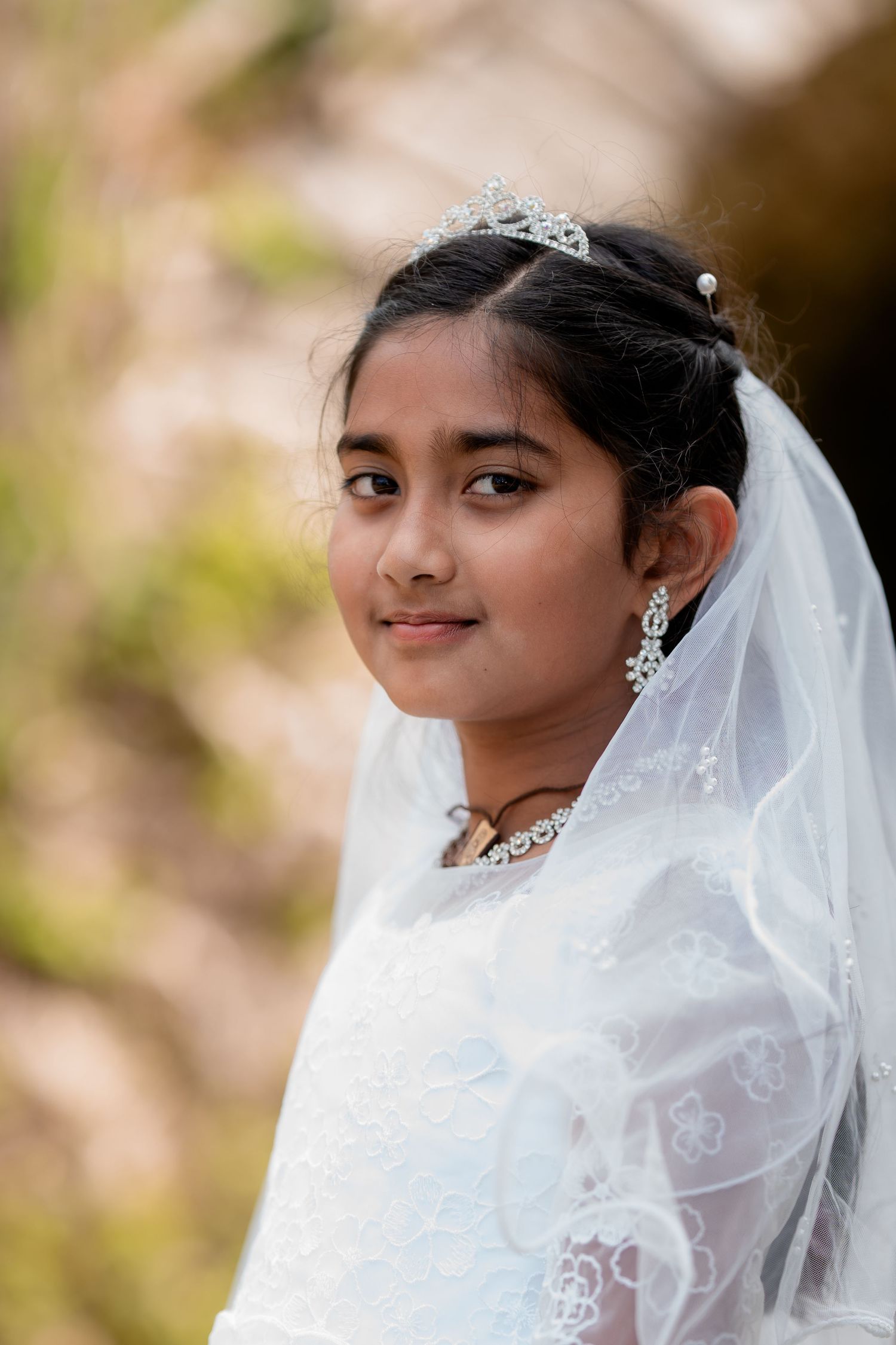 Young girl in white First Communion dress, veil, and tiara smiling outdoors.