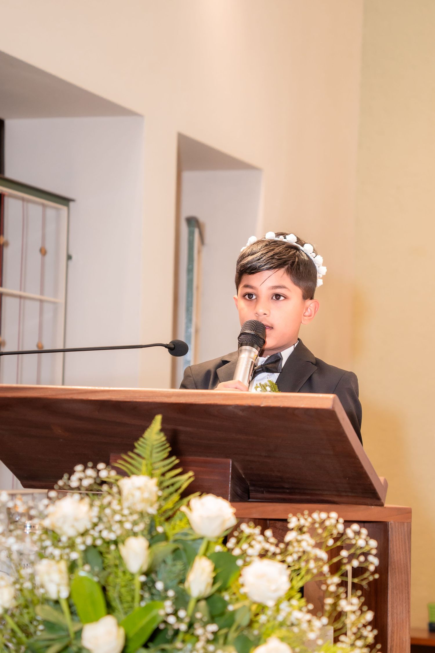 Young boy in suit speaks at podium with microphone during First Communion ceremony, white flowers in foreground.