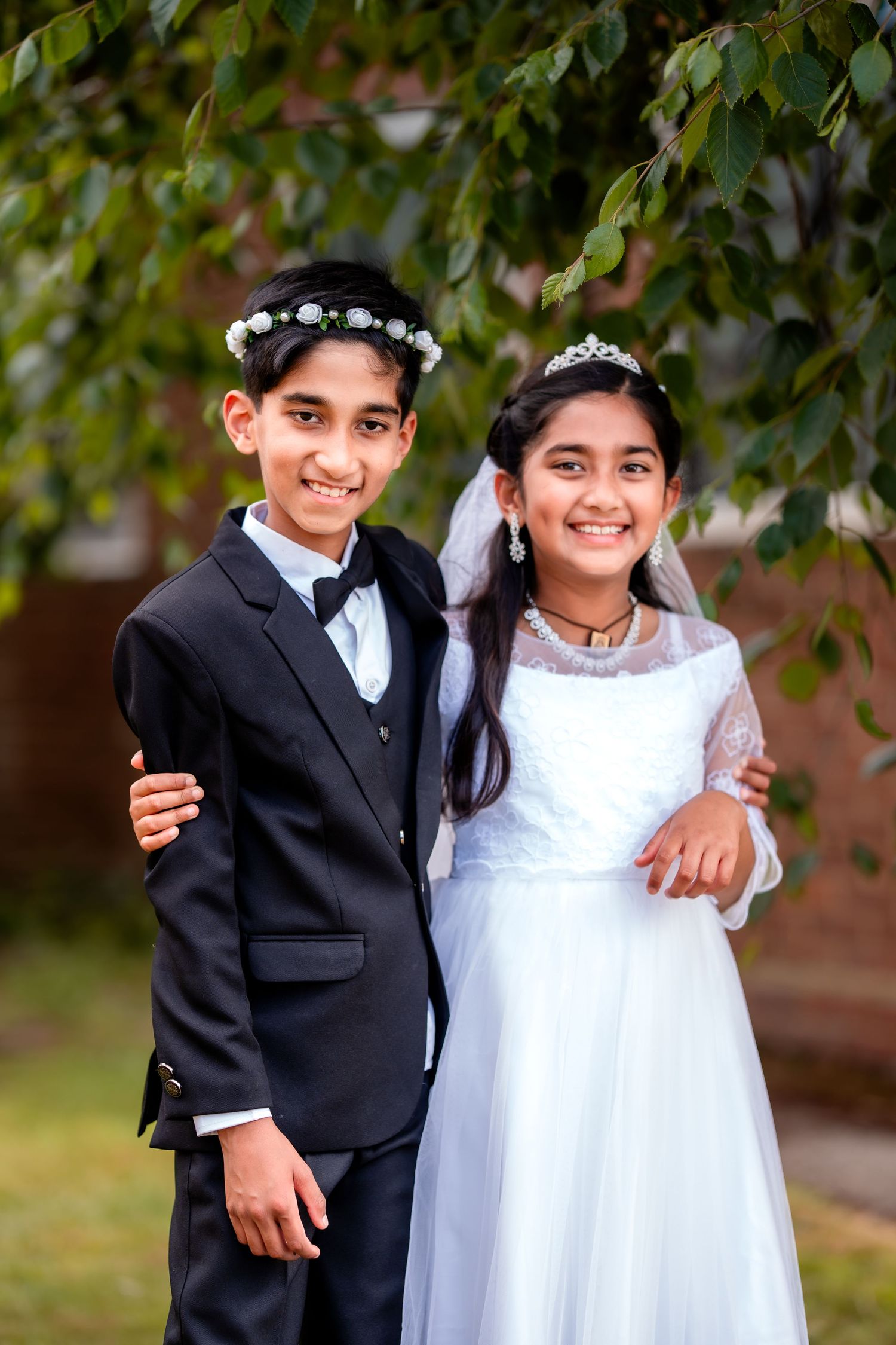 Two children in formal attire, boy in black suit and girl in white dress with veil, posing outdoors for First Communion.