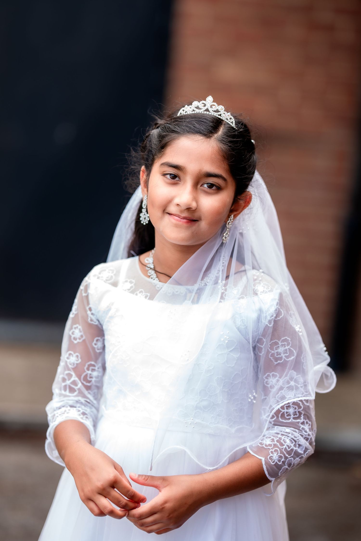 Young girl in white First Communion dress, lace veil, and tiara smiling outdoors.