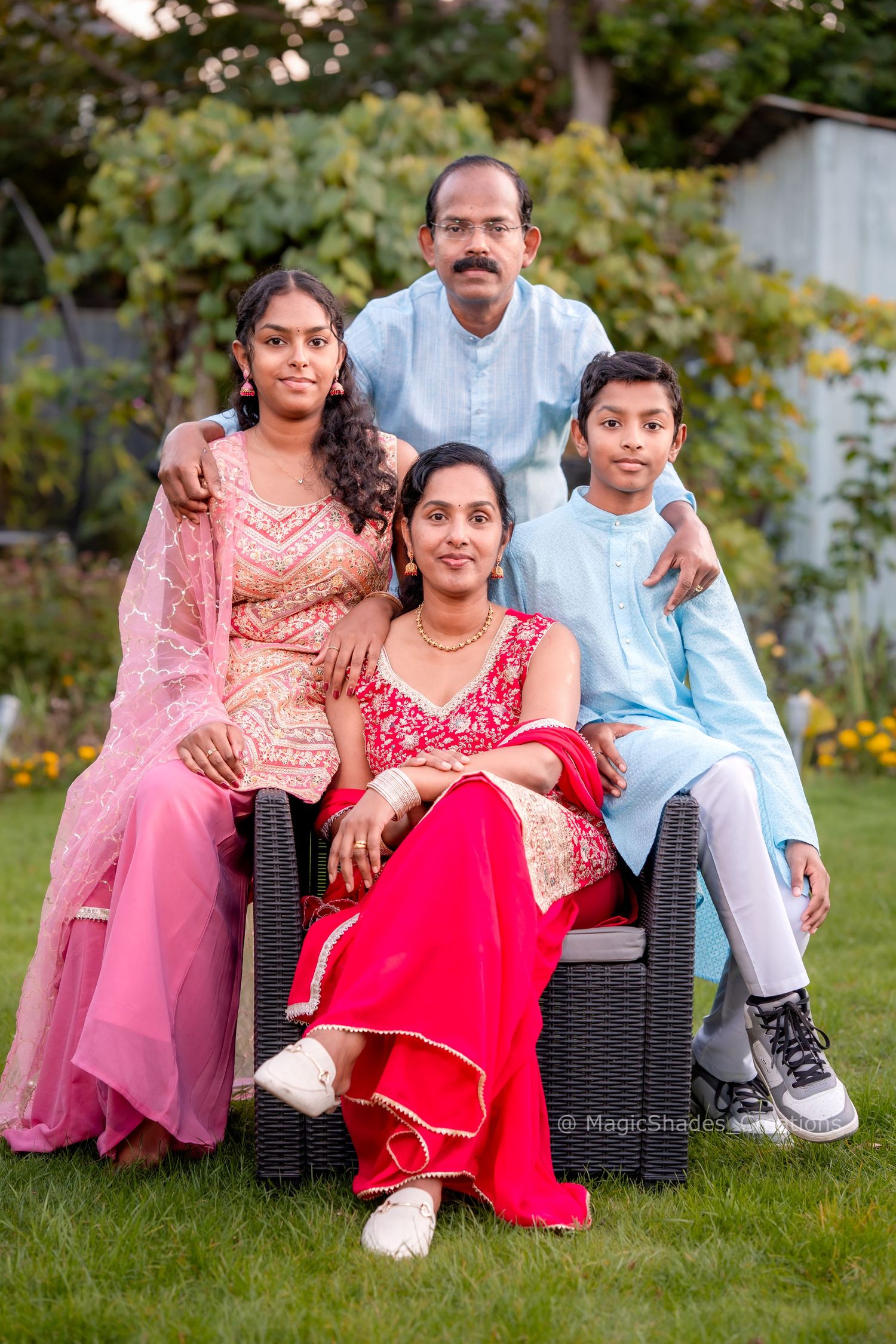 A South Asian family of four poses outdoors in colorful traditional attire, smiling together in a lush garden setting.