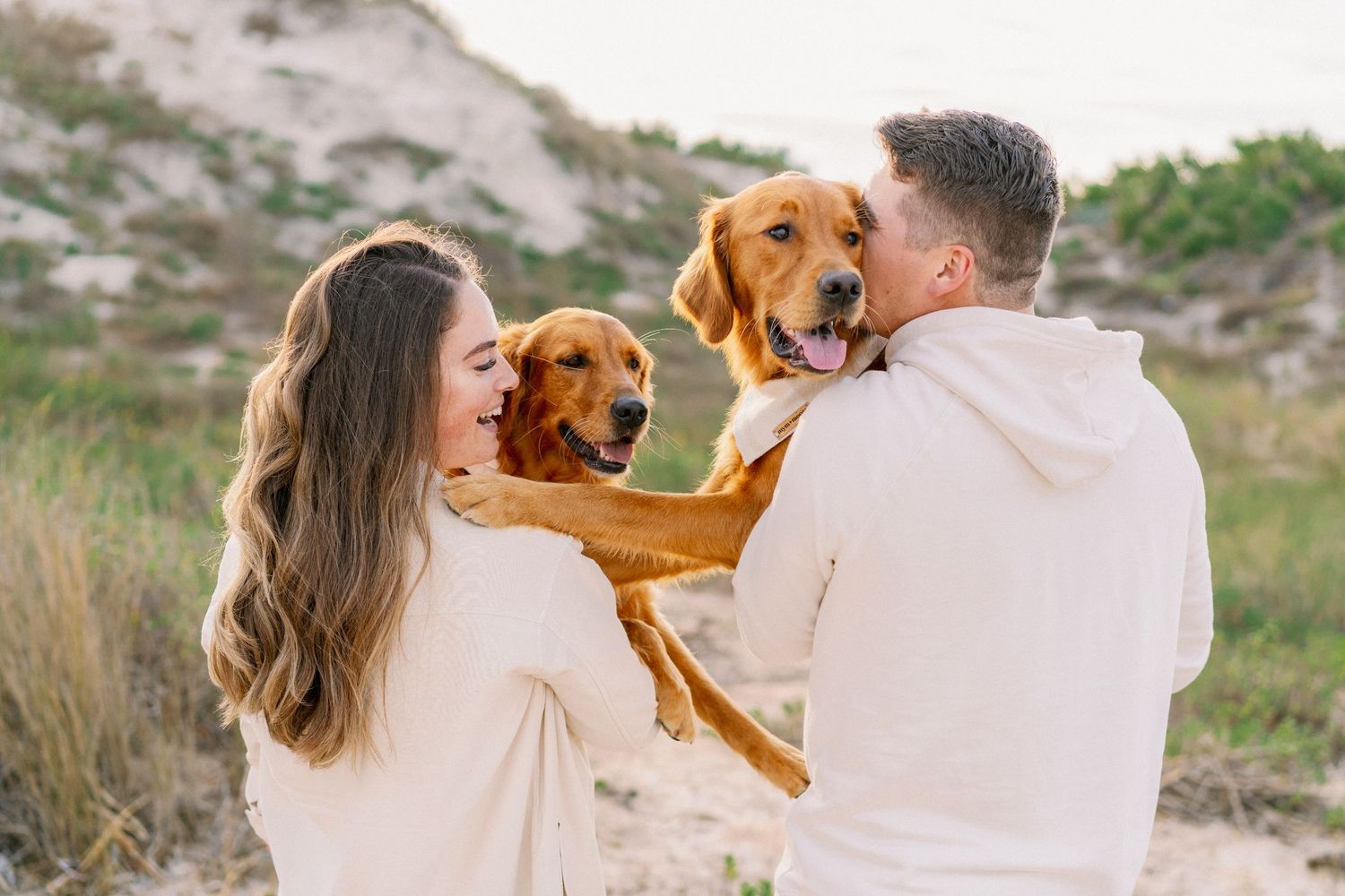A couple in white clothing walks along a beach path with their brown dog during a sunny outdoor photo session.