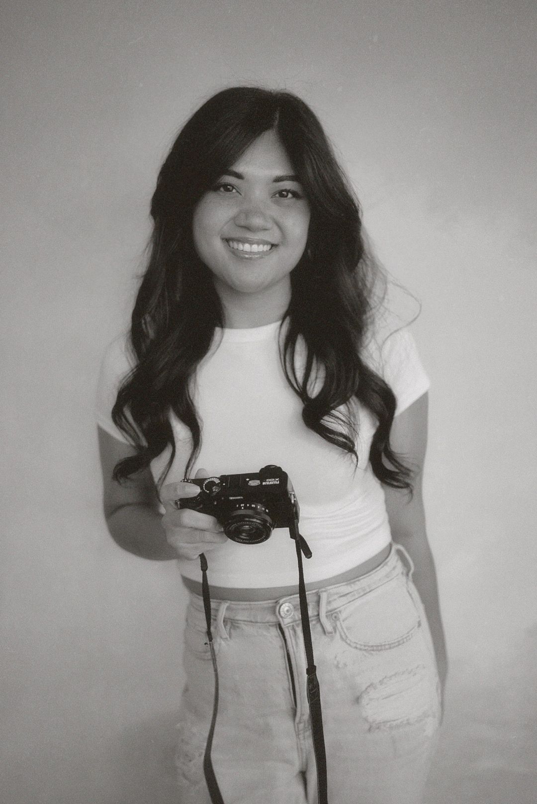 A photographer in a white t-shirt and light jeans holds a vintage camera while posing against a gray background.